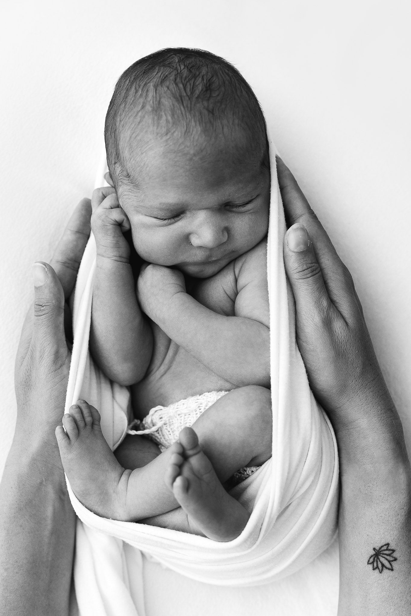 Black and white photo of a newborn baby cradled in adult hands, wrapped in white fabric, peacefully resting.