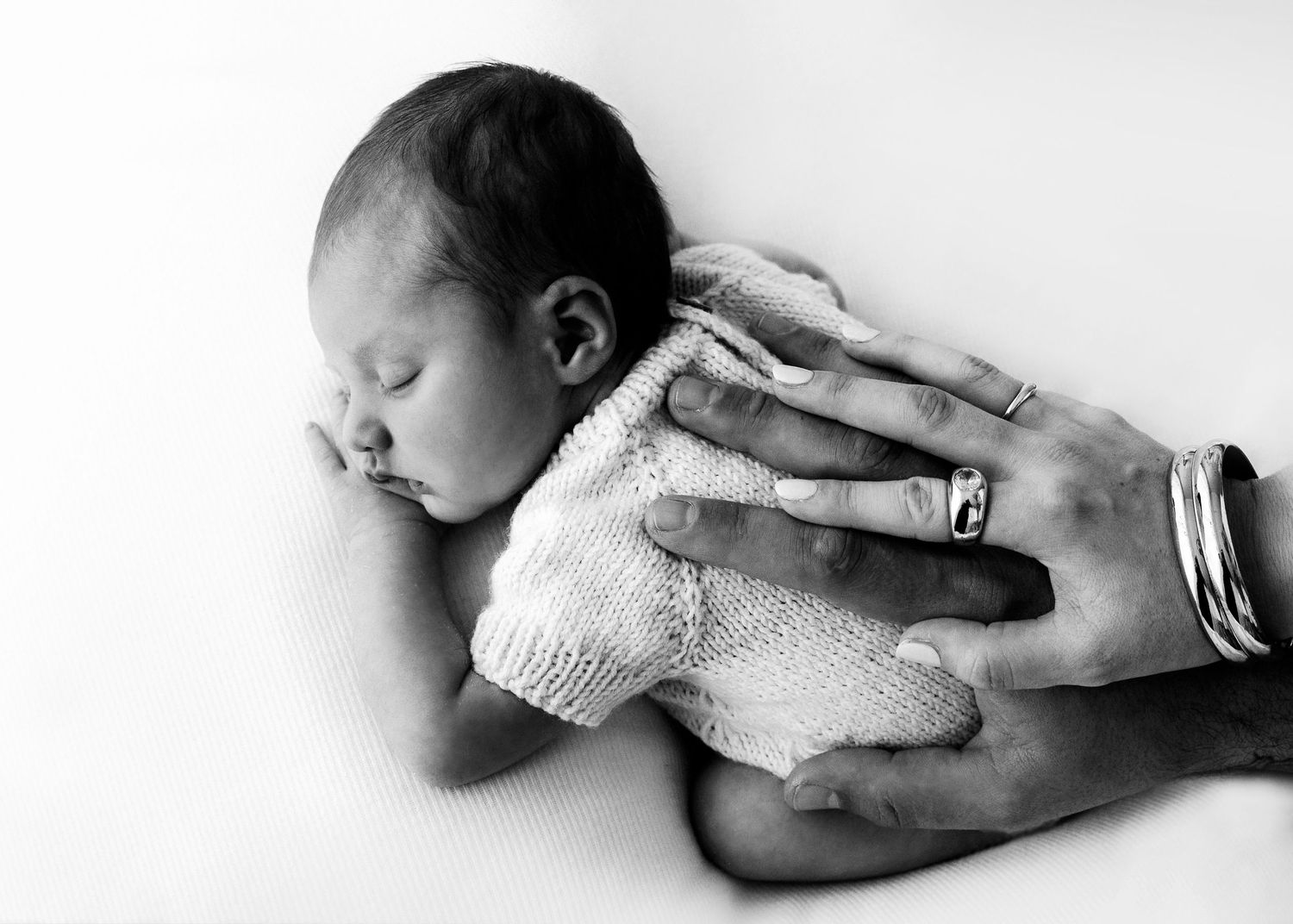 Black and white photo of a sleeping newborn baby cradled in adult hands, wearing a knit outfit.