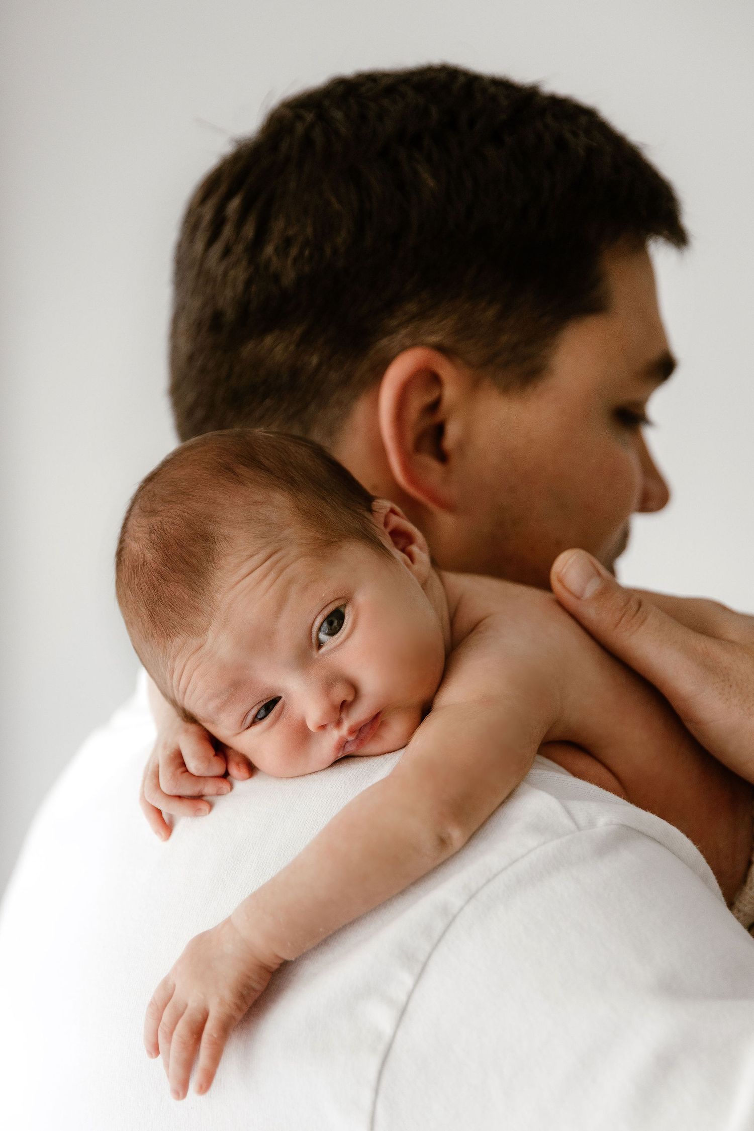 Newborn baby resting on fathers shoulder looking at camera during professional newborn photo session.