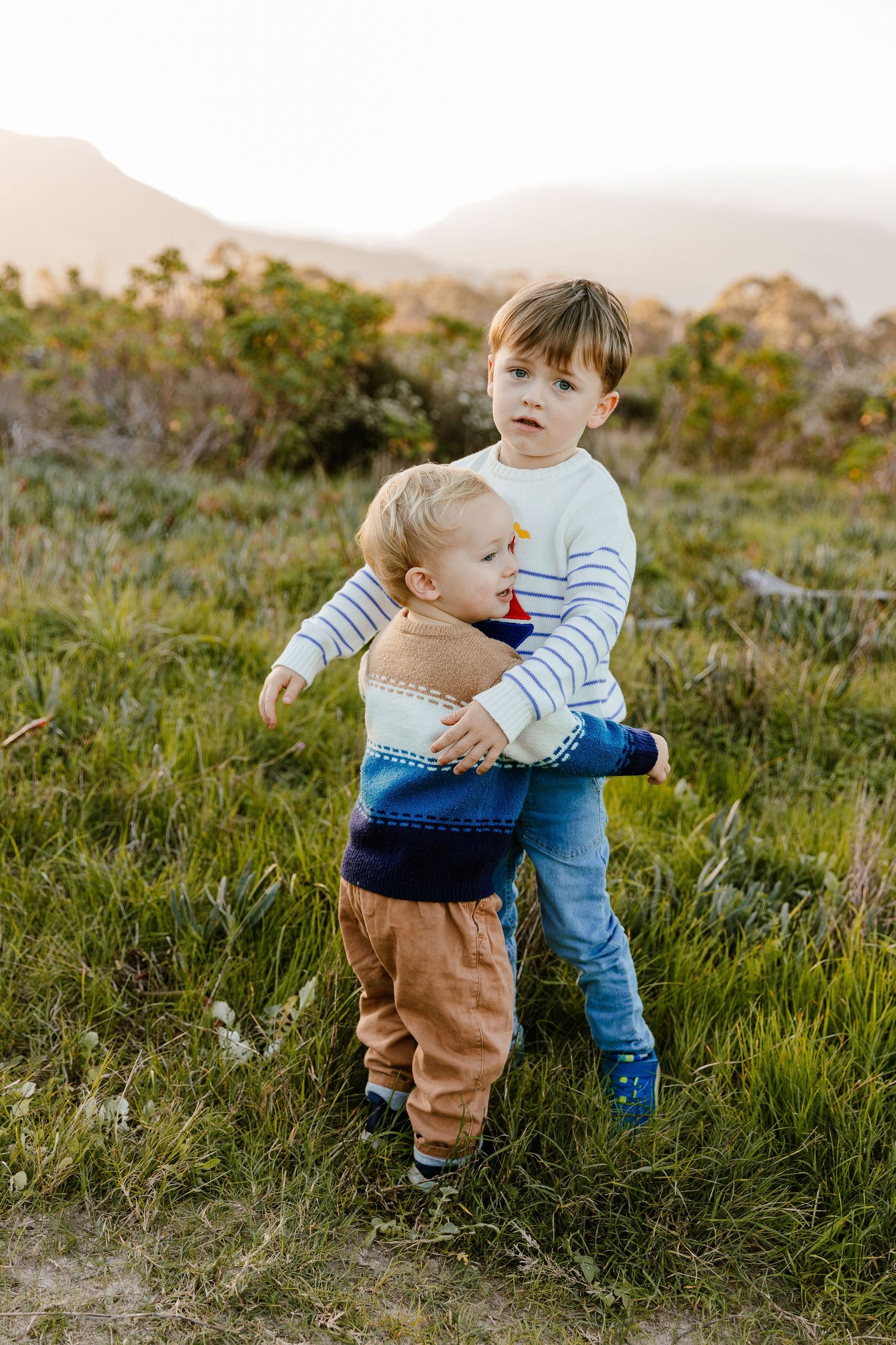 Two young brothers hugging each other in a green grassy field with mountains in the background.