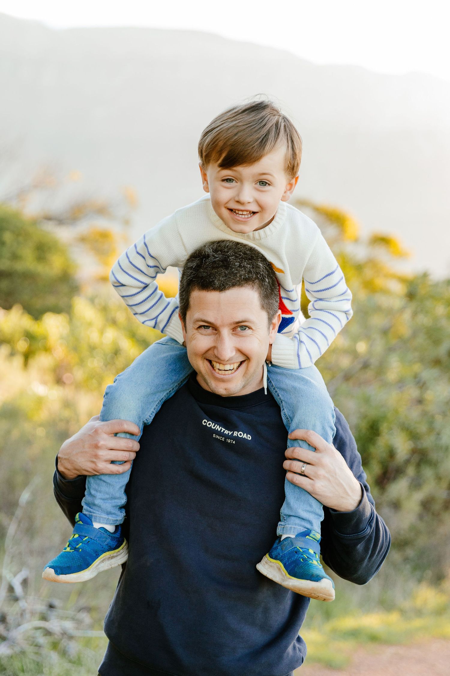 Smiling father carrying young boy on shoulders in sunny outdoor nature setting.
