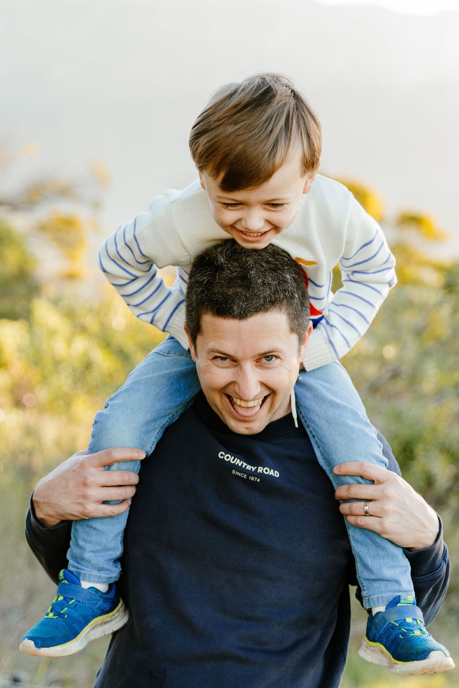 Laughing father and young son sharing a joyful piggyback ride outdoors in golden afternoon light.