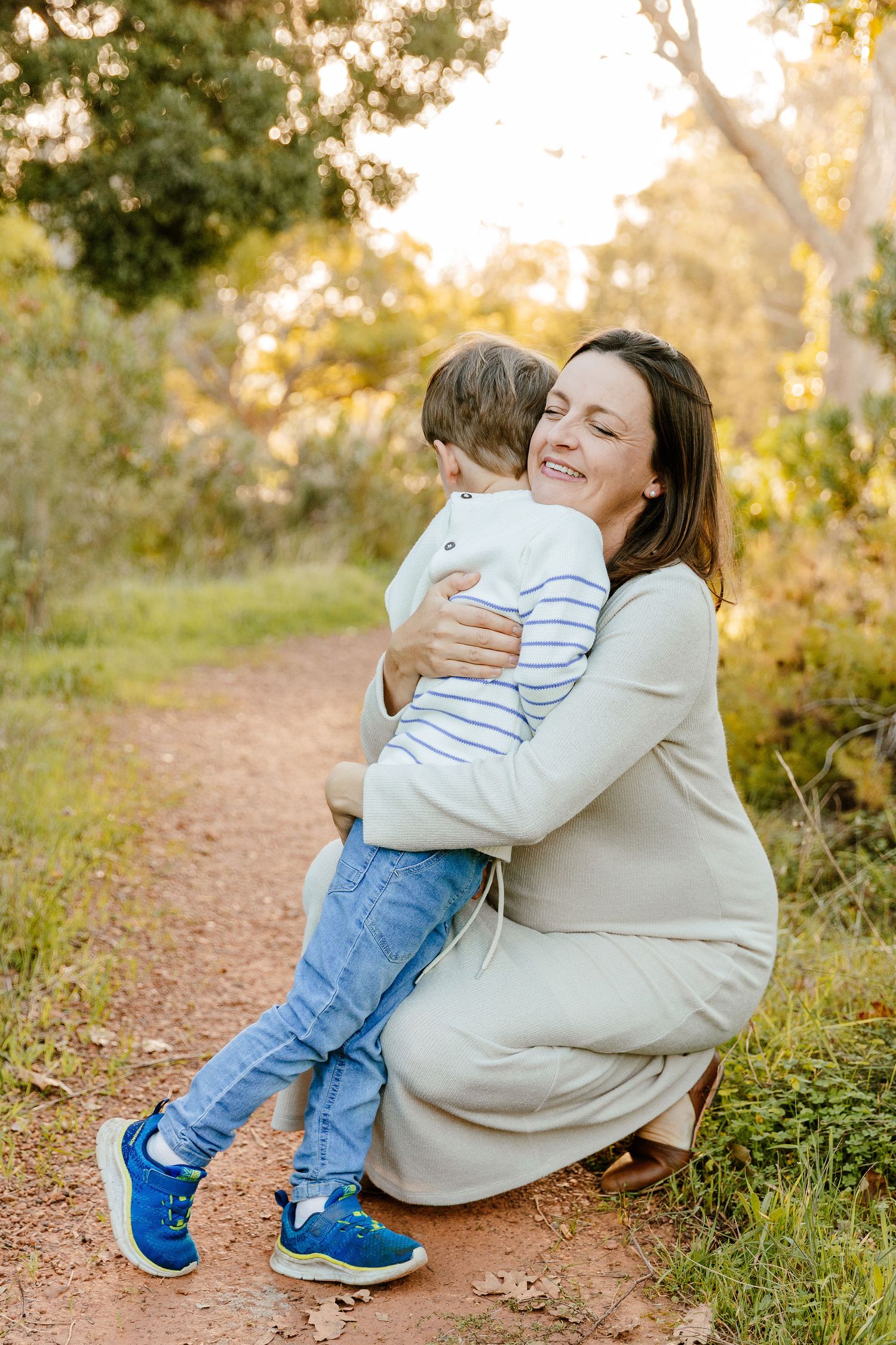 Pregnant mother in white dress hugging young son on a sunlit nature trail.