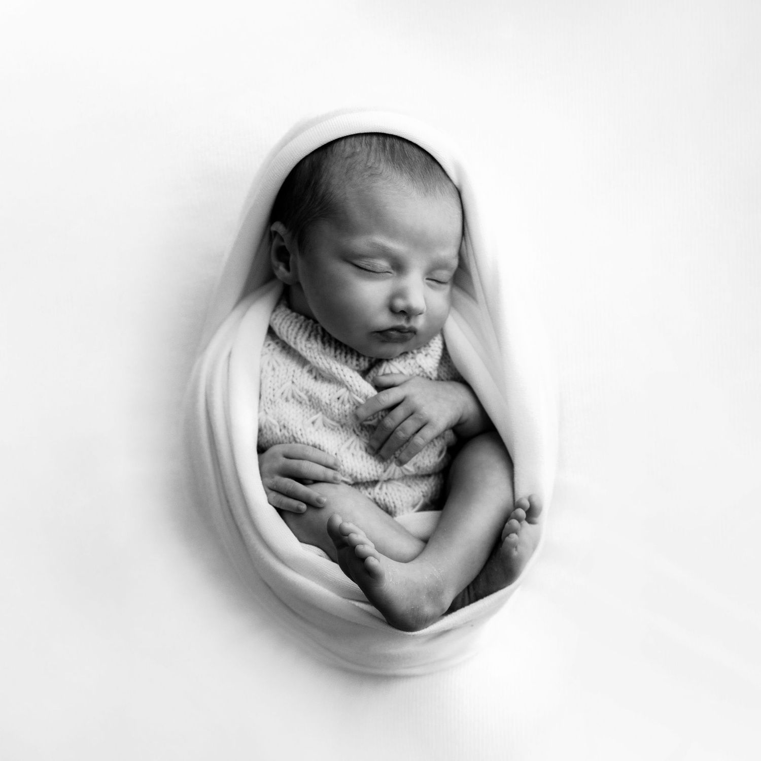 Black and white overhead portrait of newborn baby peacefully sleeping swaddled in a soft white wrap.