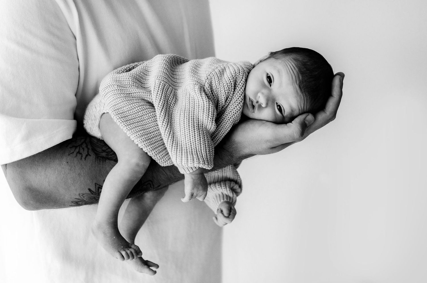 Father cradling sleeping newborn baby on forearm in black and white photo.
