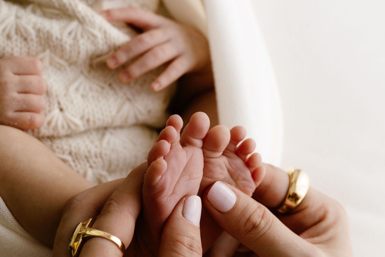 Close-up of newborn baby feet cradled in mother's hands wearing gold rings.