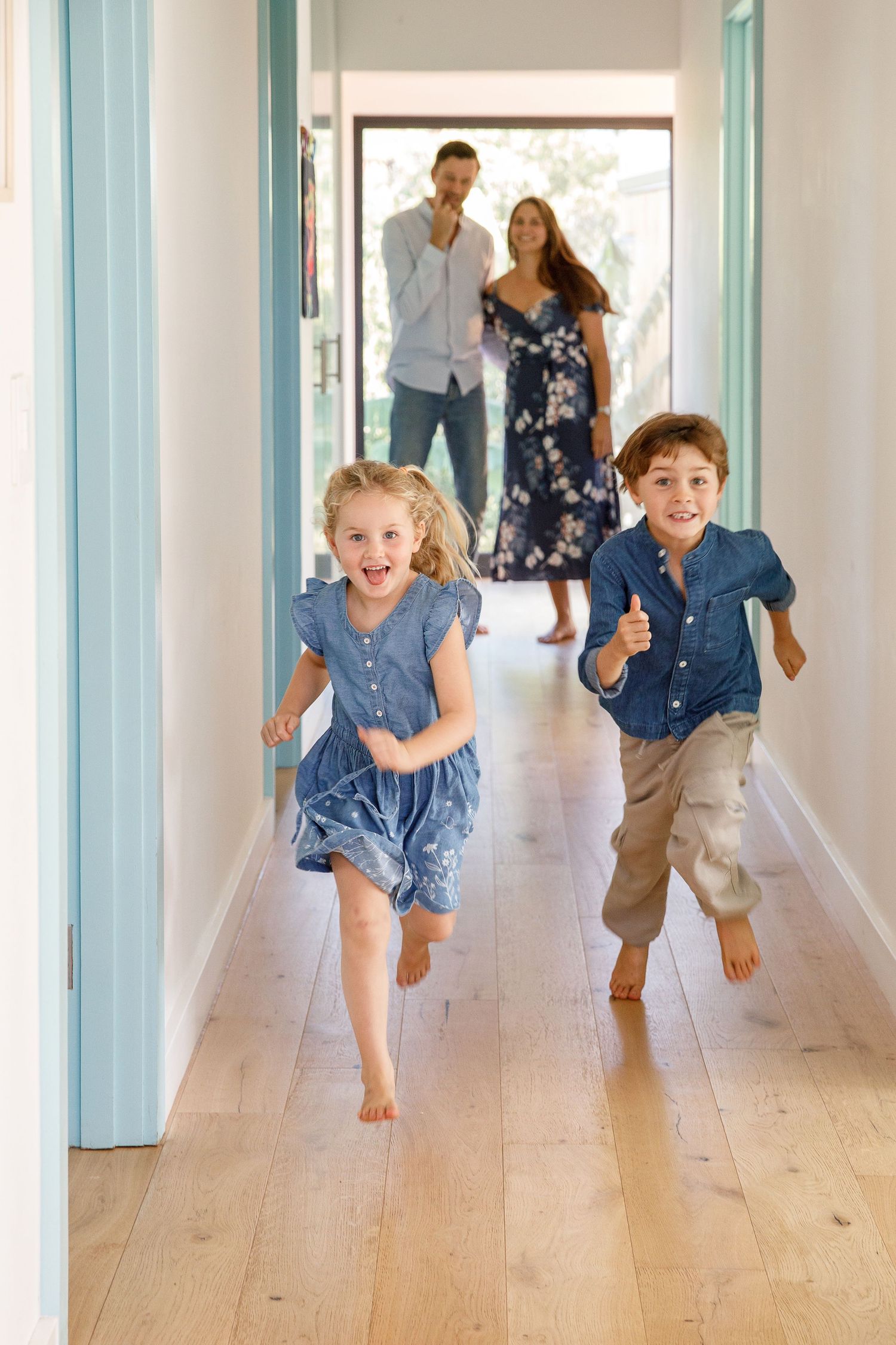 Two excited children running barefoot down a hallway while their parents watch smiling in the background.