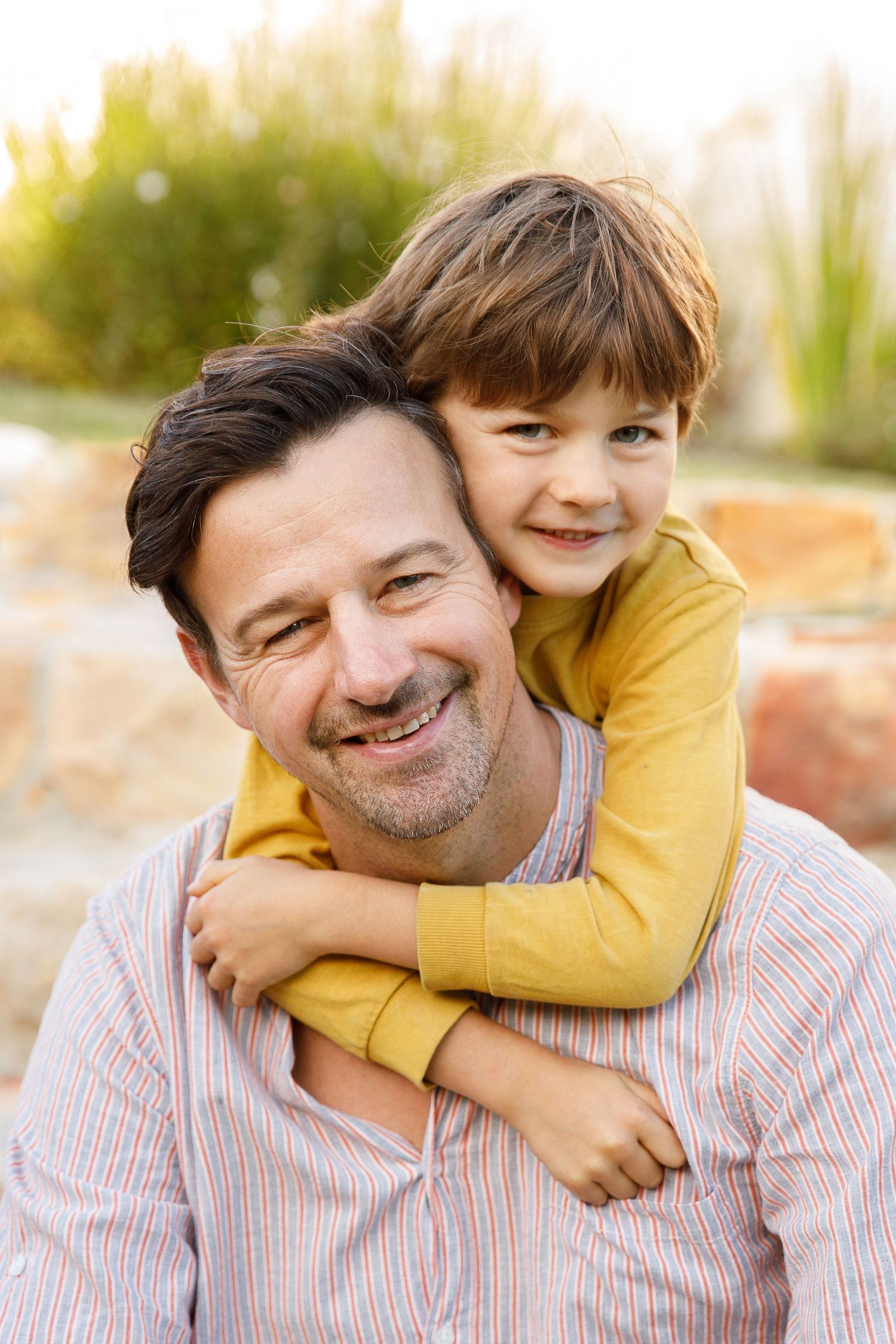 Smiling father with young son hugging him from behind outdoors in warm sunlight.