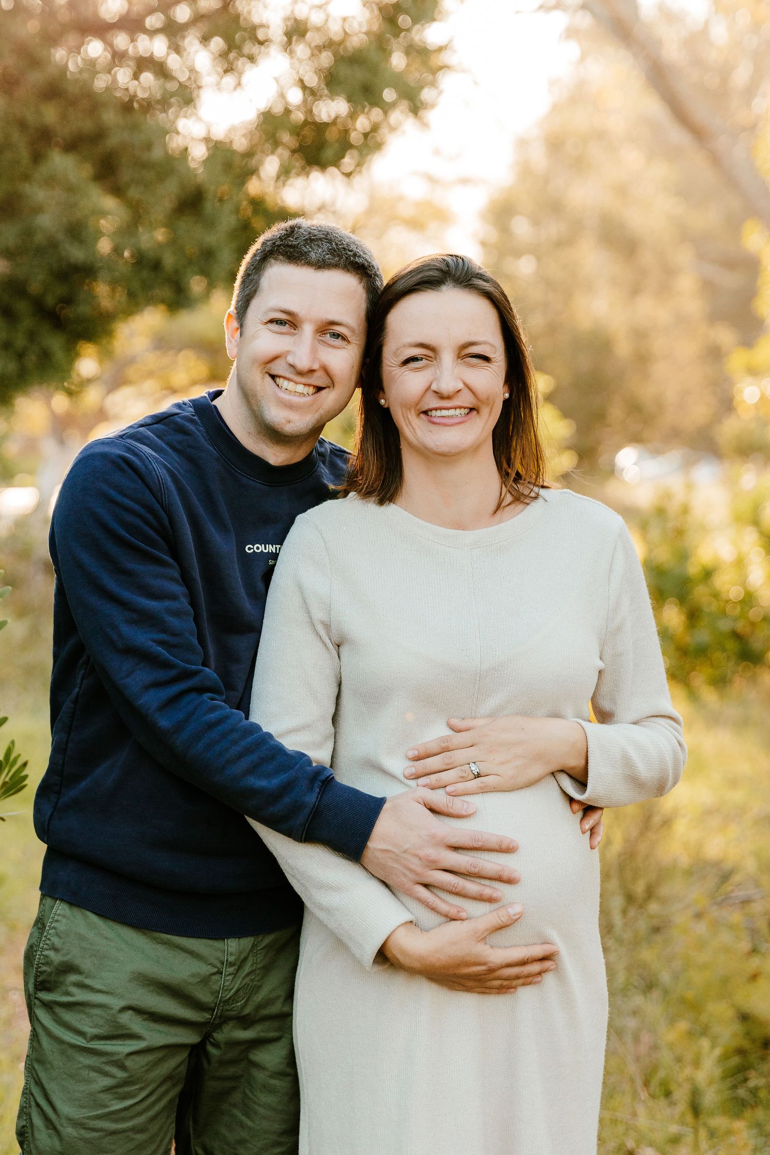 Happy couple posing outdoors, woman in white dress cradling baby bump, man in navy sweater embracing her warmly.