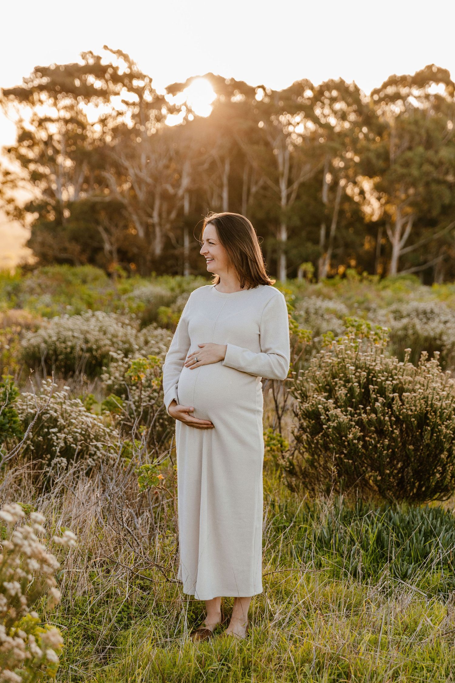 Smiling pregnant woman in cream dress cradling baby bump at golden hour in lush green field.