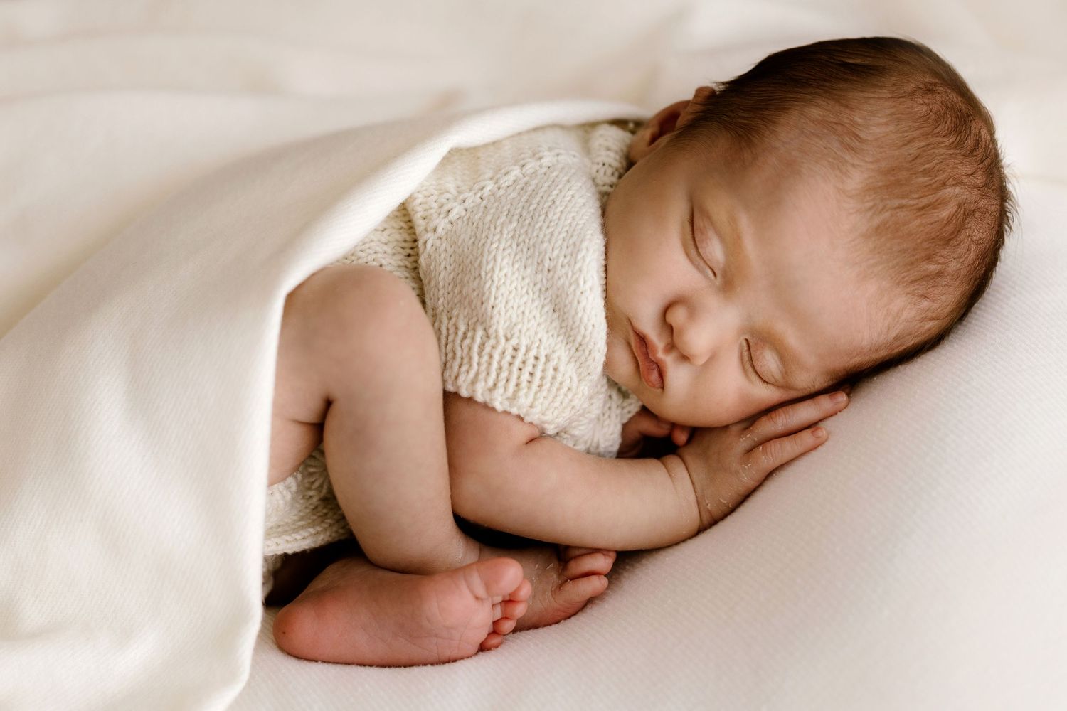 Peaceful newborn baby sleeping curled up under a soft white blanket in a cozy studio setting.