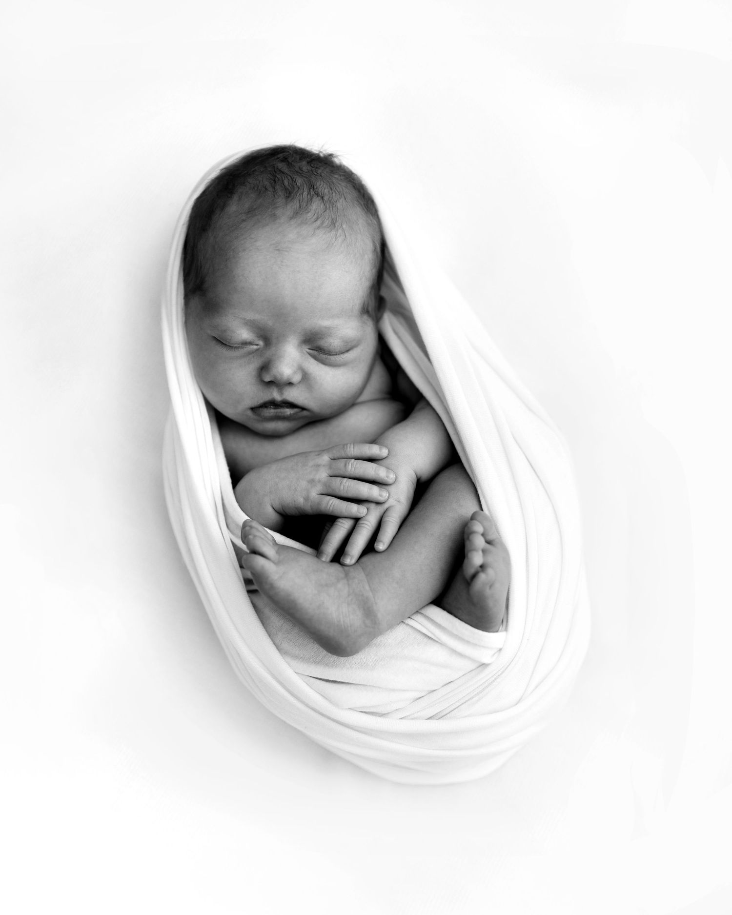 Black and white photo of a sleeping newborn baby wrapped in white cloth, curled in fetal position.