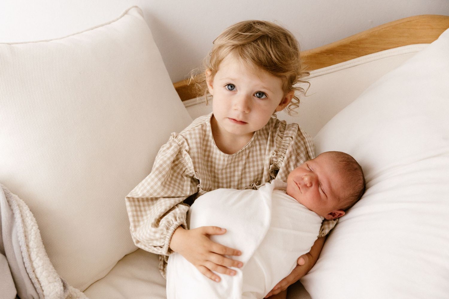 Toddler with blonde hair gently holds a sleeping newborn baby wrapped in white, sitting on a white bed.
