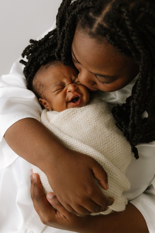 A mother with braided hair tenderly kisses her laughing newborn baby wrapped in a white knit blanket.