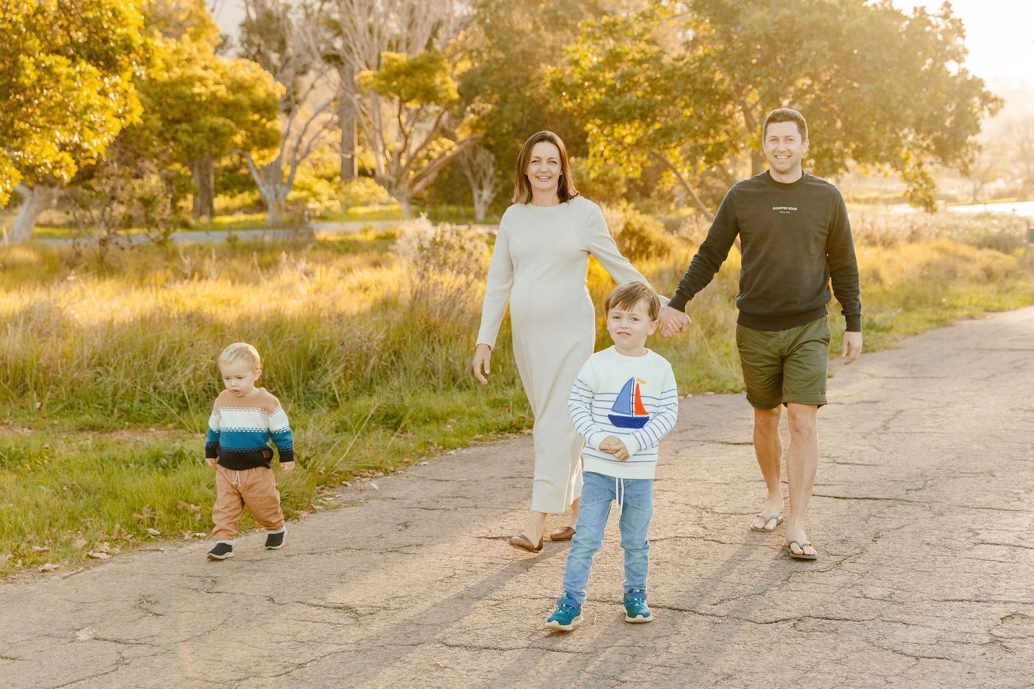 Family of four walking on a sunlit path, pregnant mother in white dress with toddler and young child at golden hour.