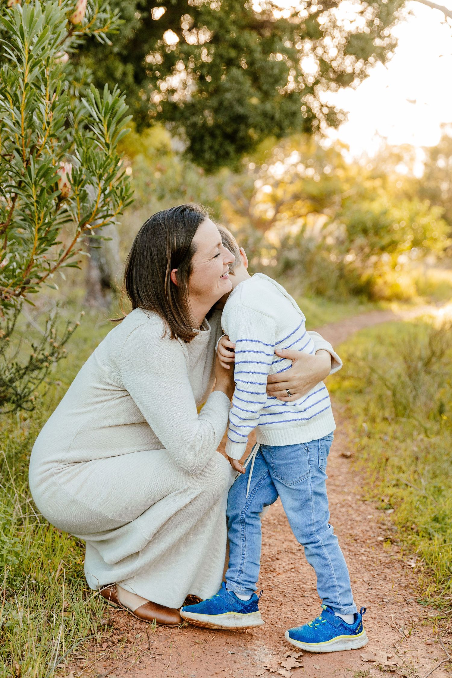 Pregnant woman in cream dress crouching to hug smiling young boy on a nature trail.