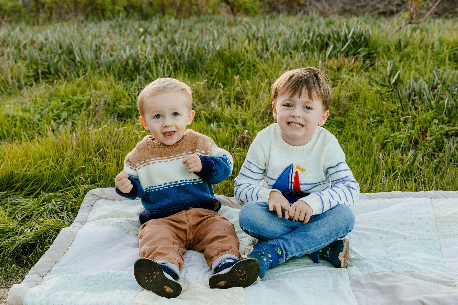 Two young brothers sitting on a blanket outdoors, wearing cozy sweaters, smiling at the camera in a grassy field.