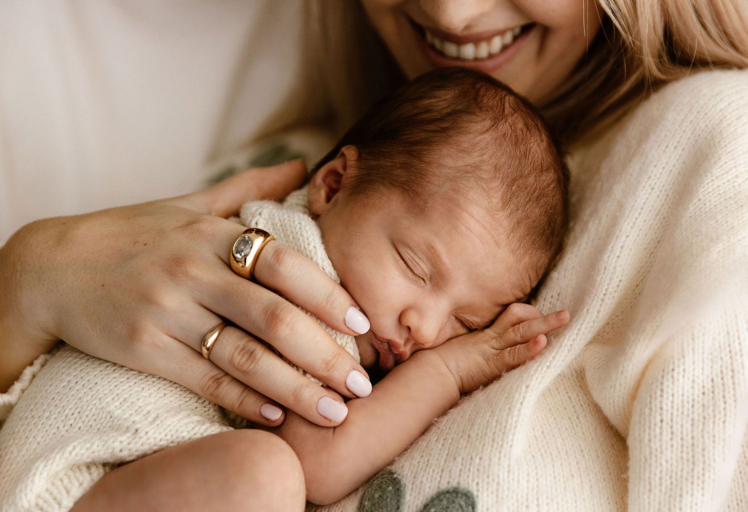 A smiling mother gently holds her peaceful sleeping newborn baby wrapped in a soft white blanket.