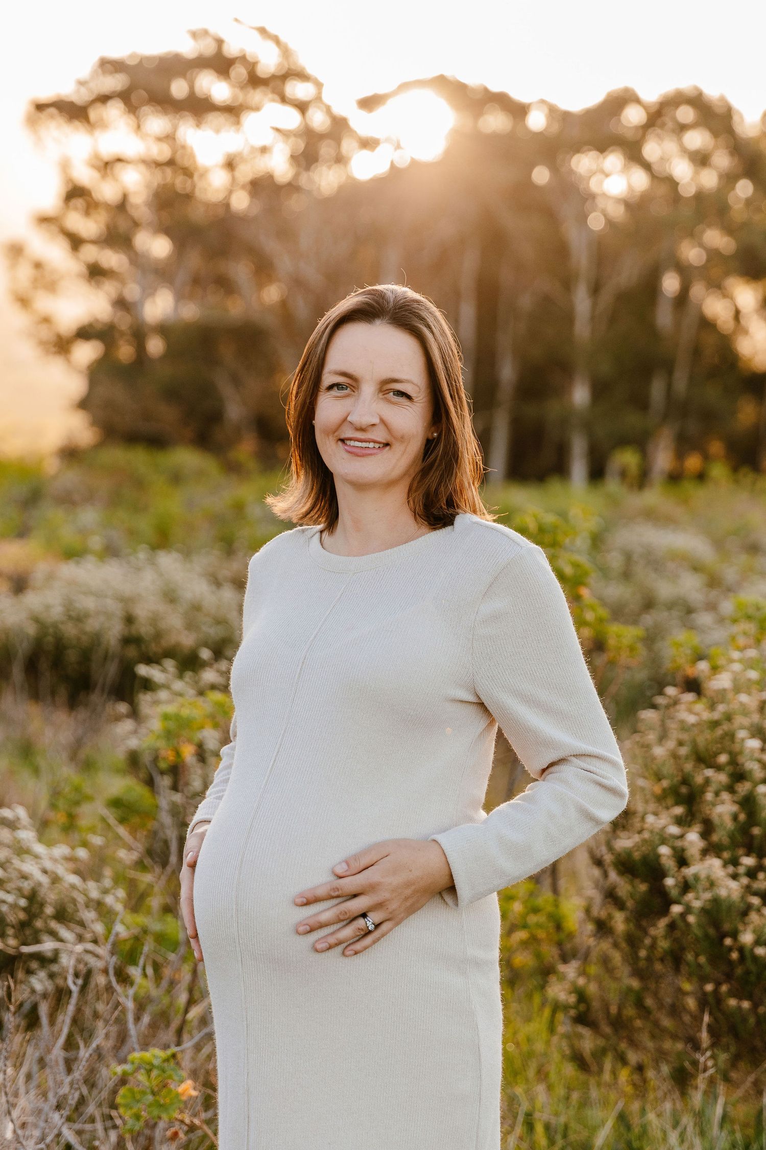 Smiling pregnant woman in white dress cradling baby bump at golden hour in a natural outdoor setting.