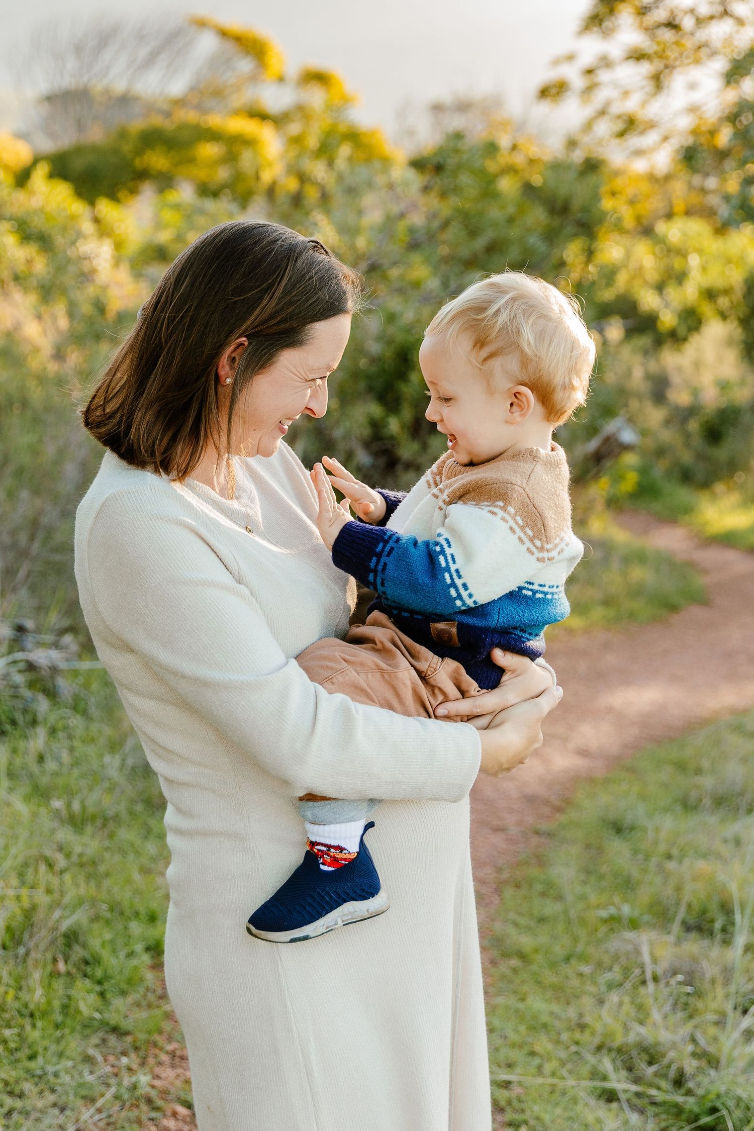 A smiling mother in a white dress holds her happy blonde toddler on a sunny outdoor path surrounded by greenery.