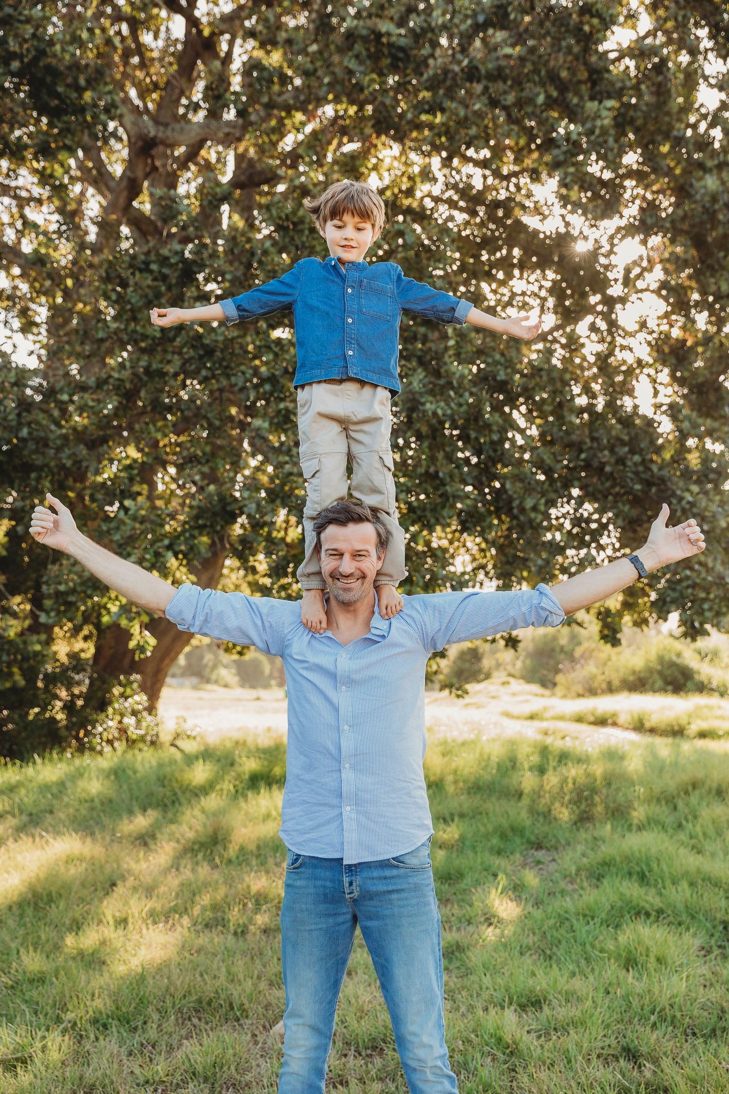 Father and son spreading arms wide outdoors, child sitting on dad's shoulders, smiling in a sunny green park.