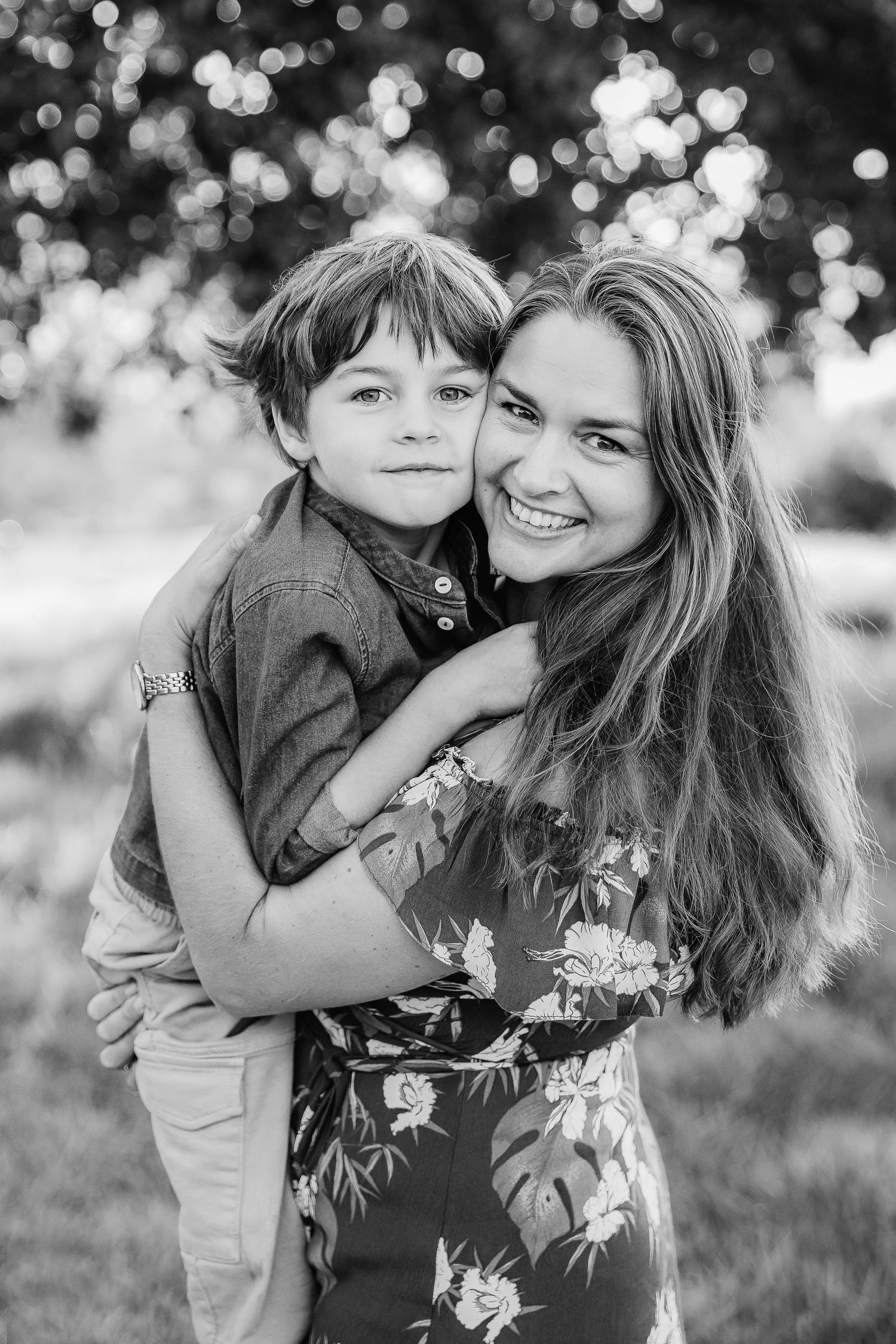 Black and white photo of a smiling mother hugging her young son outdoors among trees.