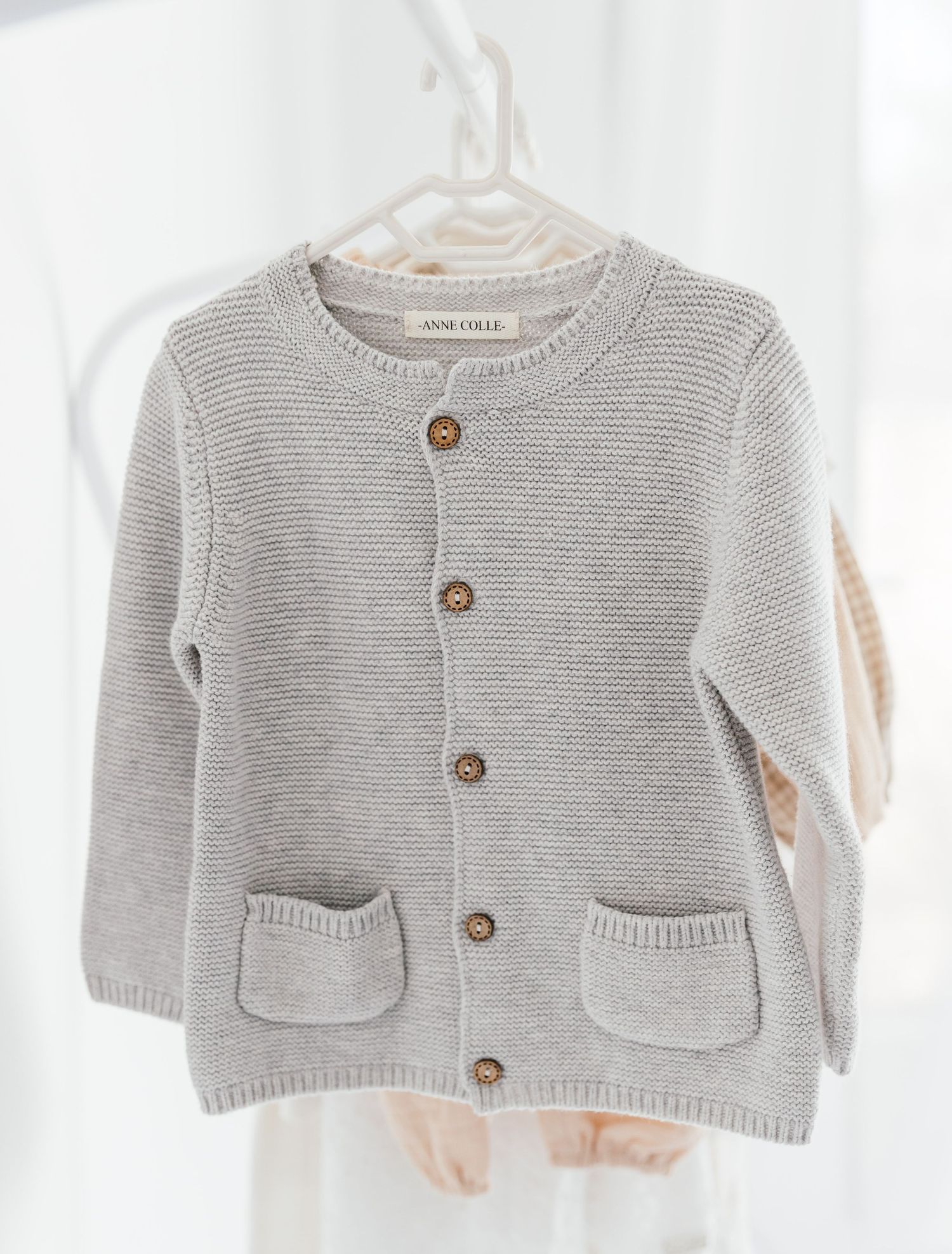 Light gray knit baby cardigan with wooden buttons and front pockets hanging on a hanger.
