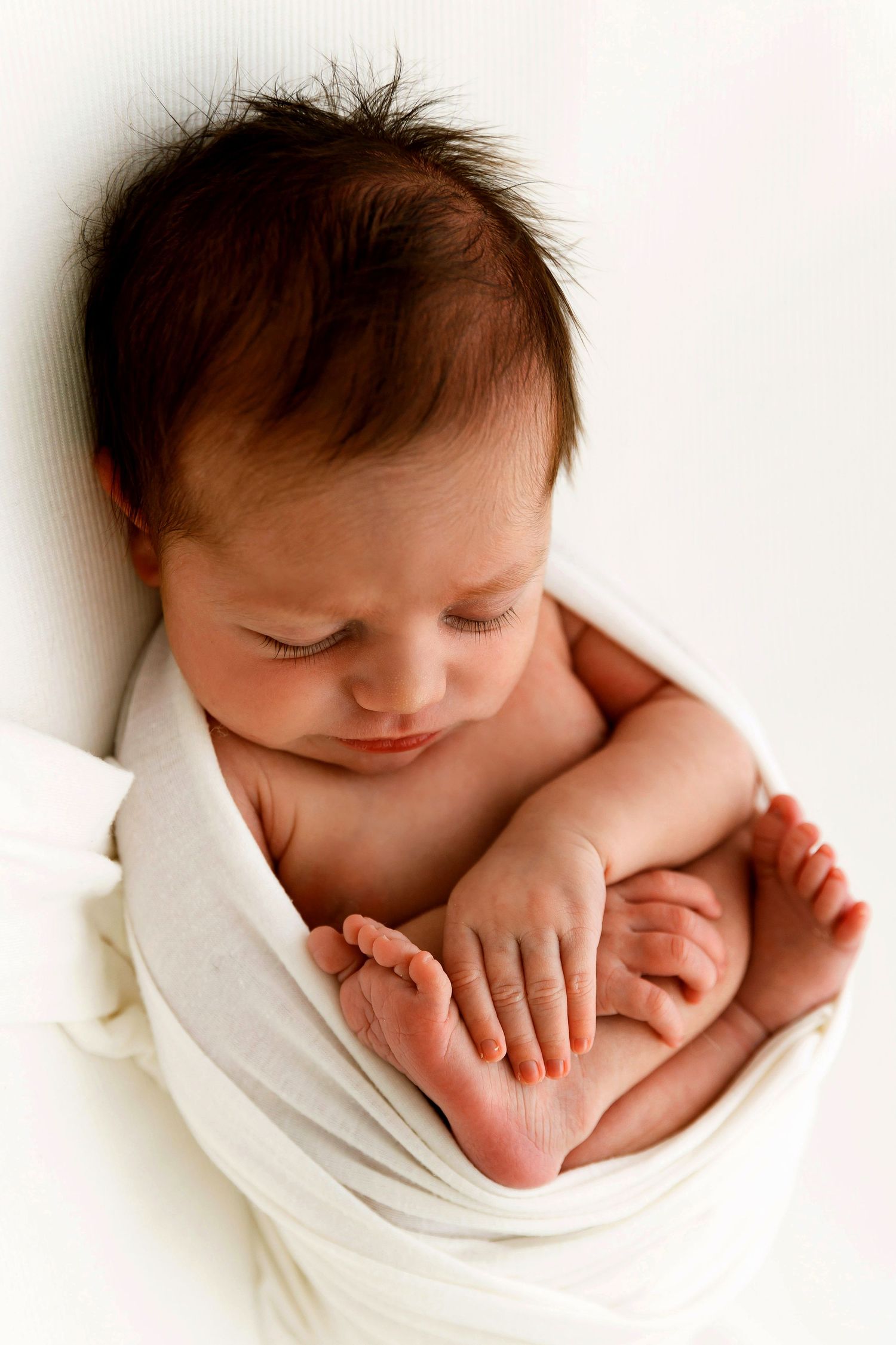 Newborn baby wrapped in white swaddle blanket, peacefully resting with tiny hands and feet visible.
