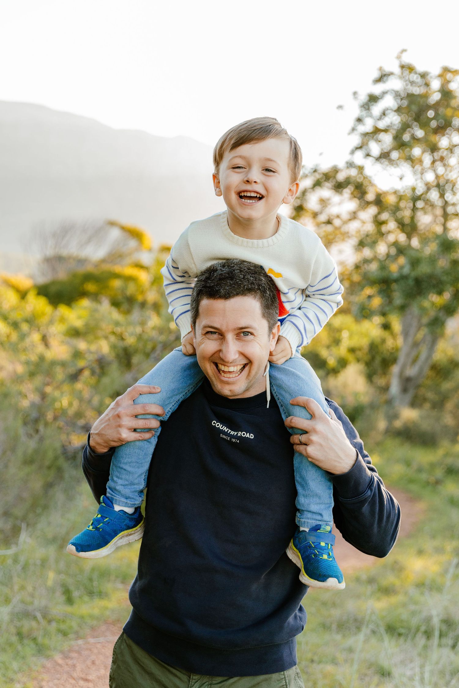 A smiling man carries a laughing young boy on his shoulders outdoors on a sunny day.