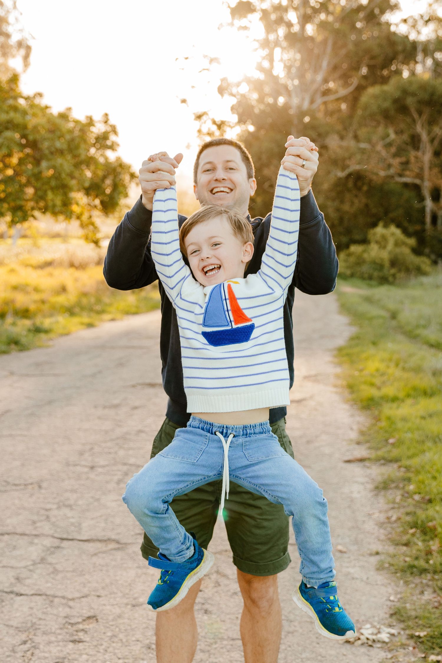 A laughing father lifts his giggling young son by the arms on a golden sunlit dirt path.