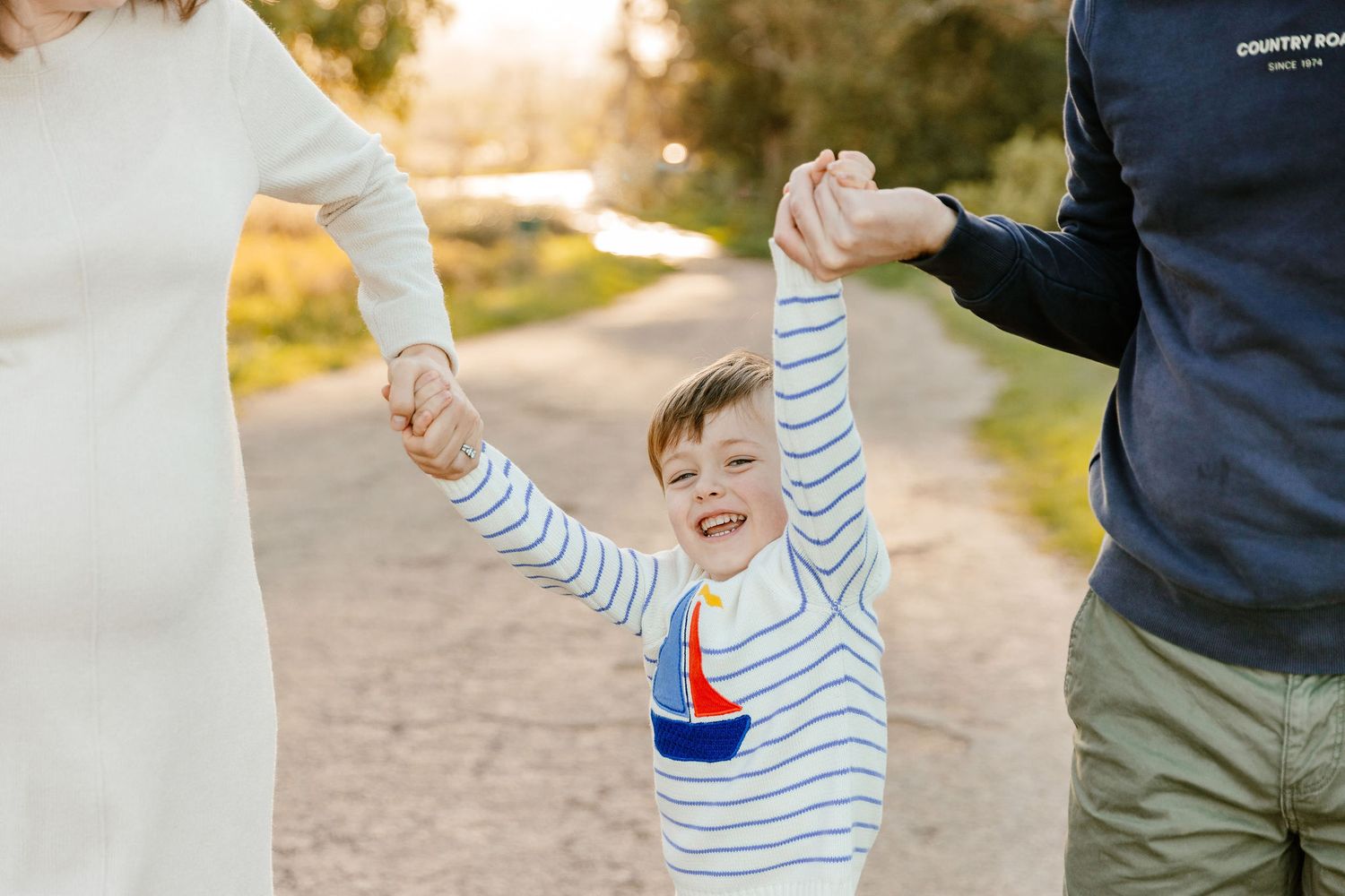 Laughing boy in striped shirt swinging between parents on a warm golden hour outdoor path.