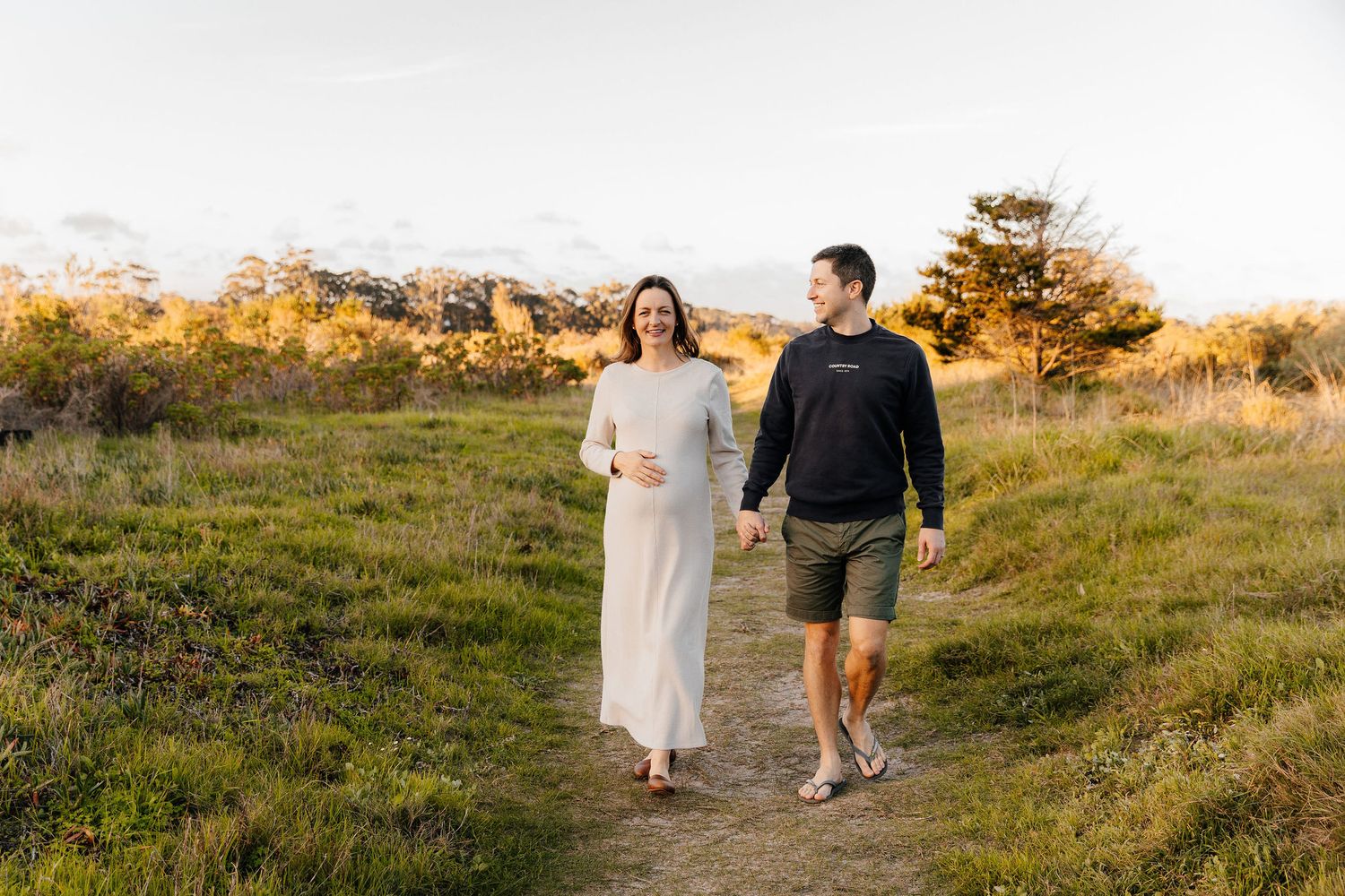 Smiling pregnant woman in a white dress walking with her husband on a sunny green nature path.
