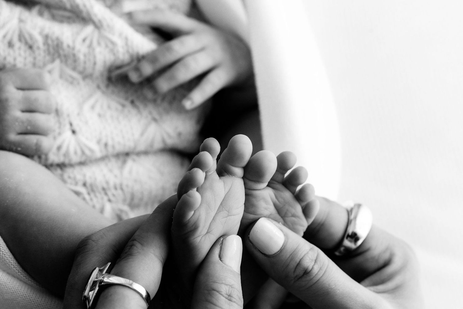 Black and white close-up of parents holding their newborn baby tiny feet with wedding rings visible.