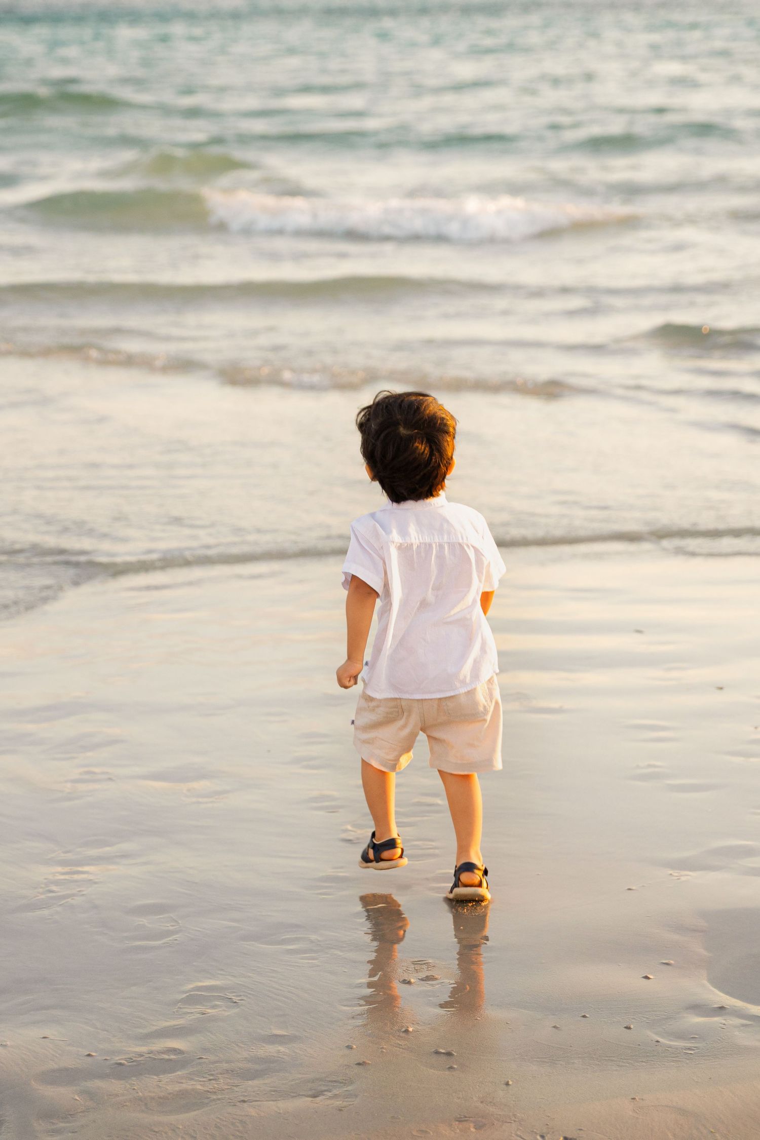 A young boy in a white shirt and shorts walks toward gentle ocean waves on a sandy beach at sunset.