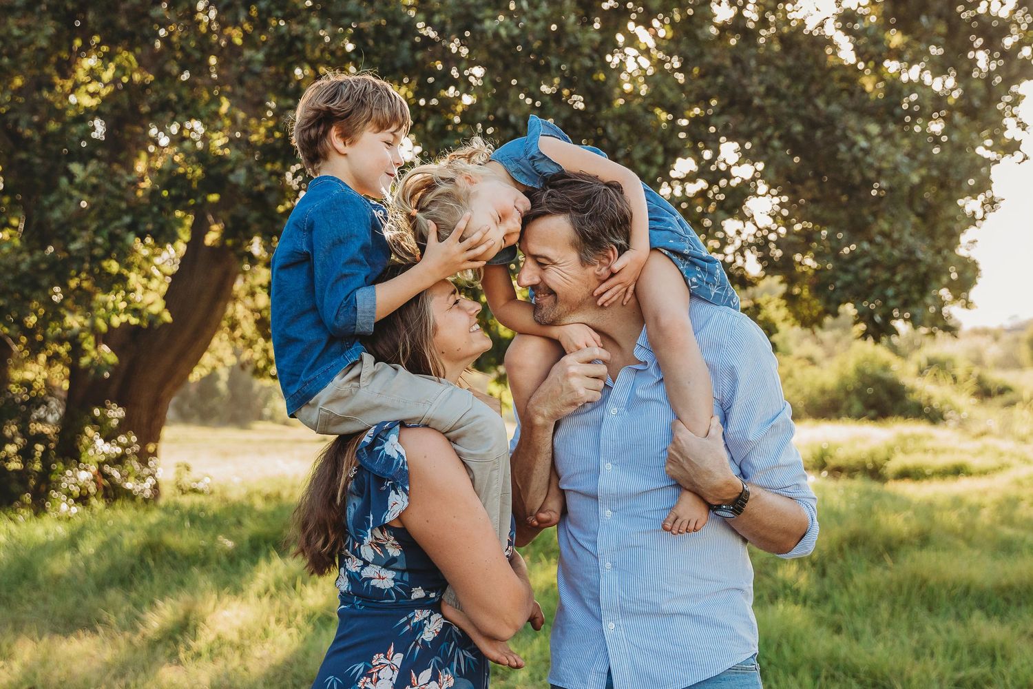 Happy family of four playing outdoors in a sunny green park, parents carrying children on shoulders while laughing together.