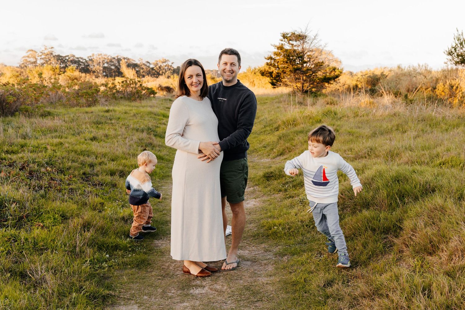 Pregnant woman in white dress poses with partner and two young children on a sunlit grassy path during golden hour.