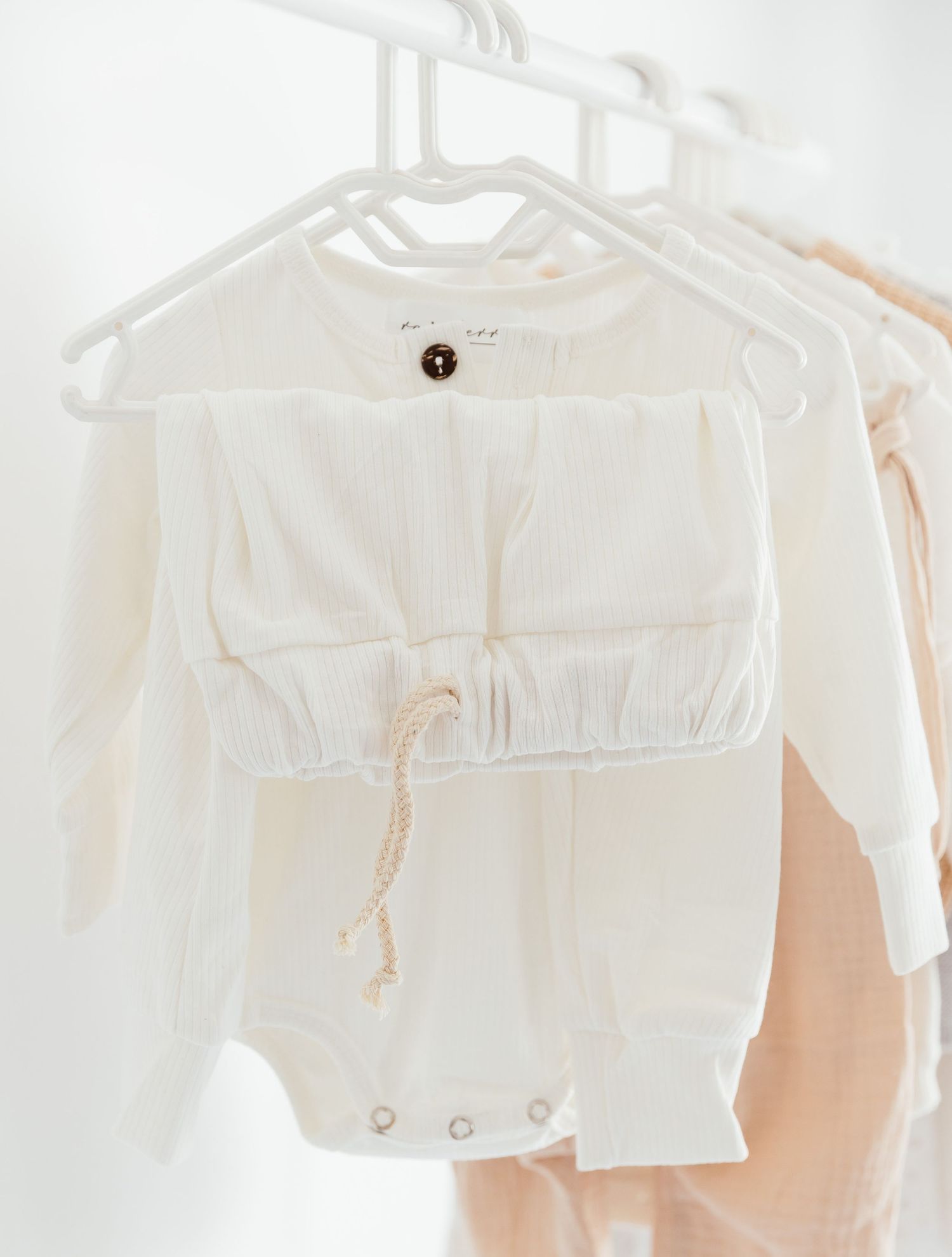 White baby romper with drawstring waist and brown button hanging on a white hanger.