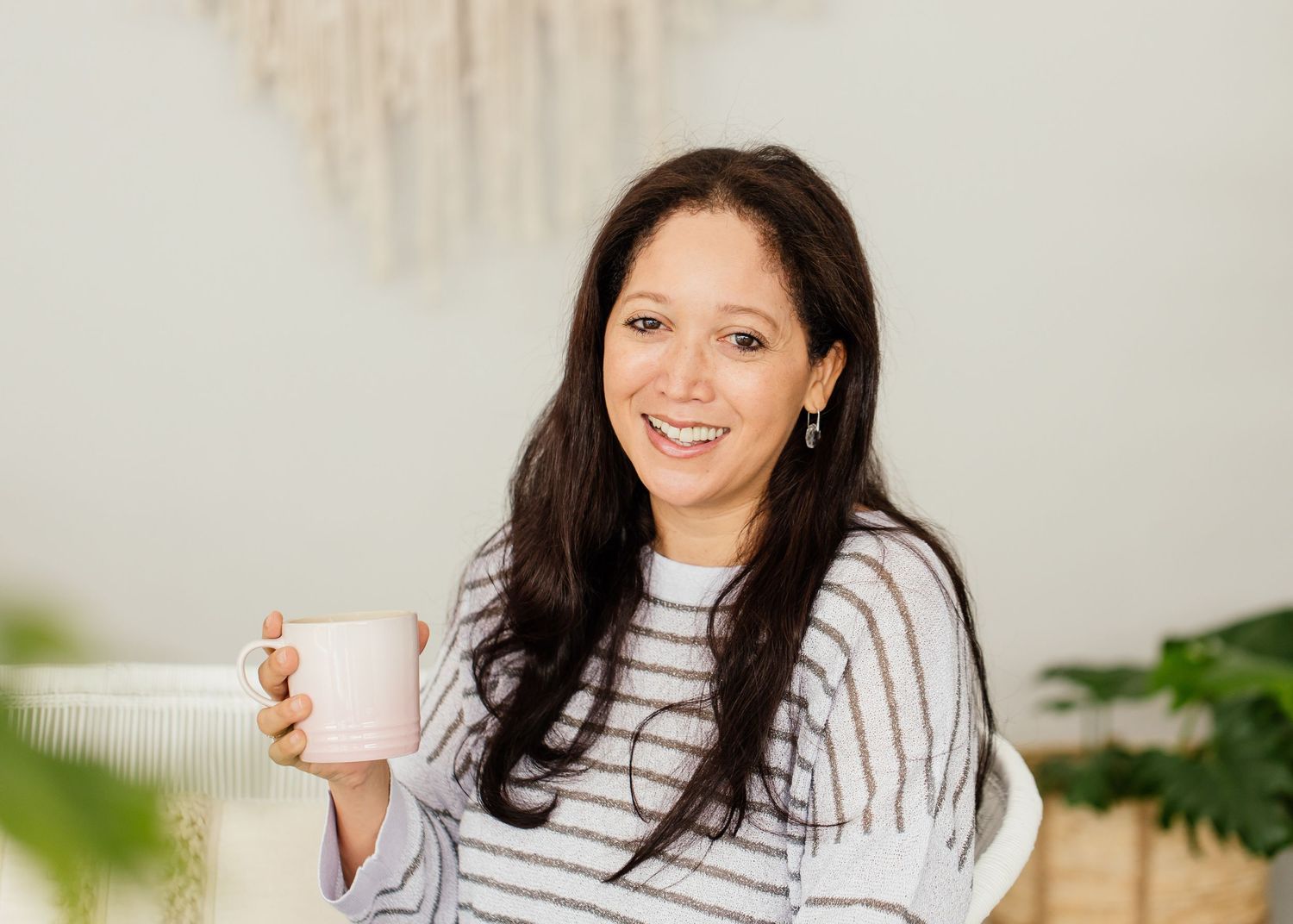 Smiling woman with dark hair holding a pink mug, wearing a striped sweater in a bright, cozy home setting.