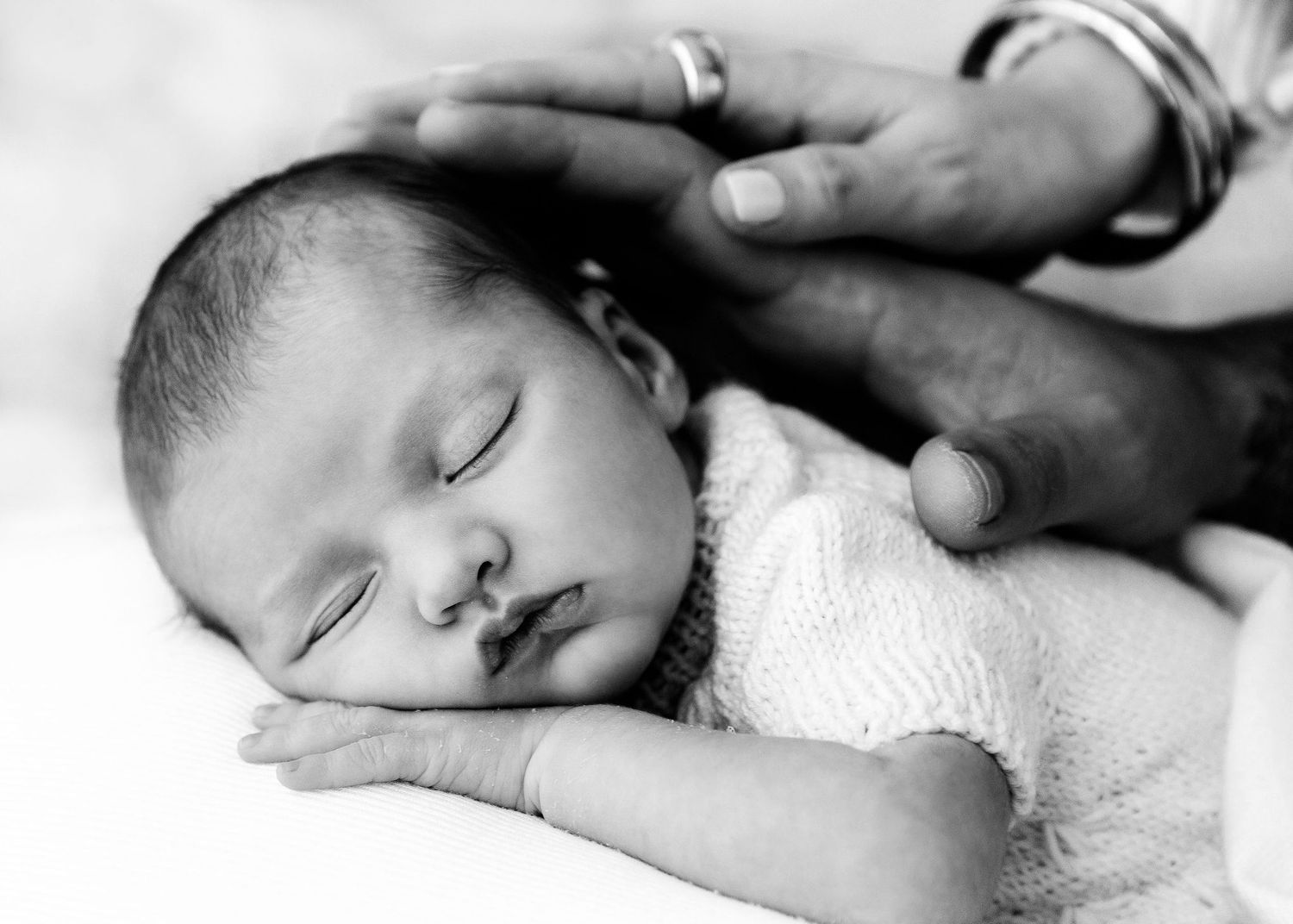 Black and white image of peaceful sleeping newborn baby with parent's gentle hand on head.