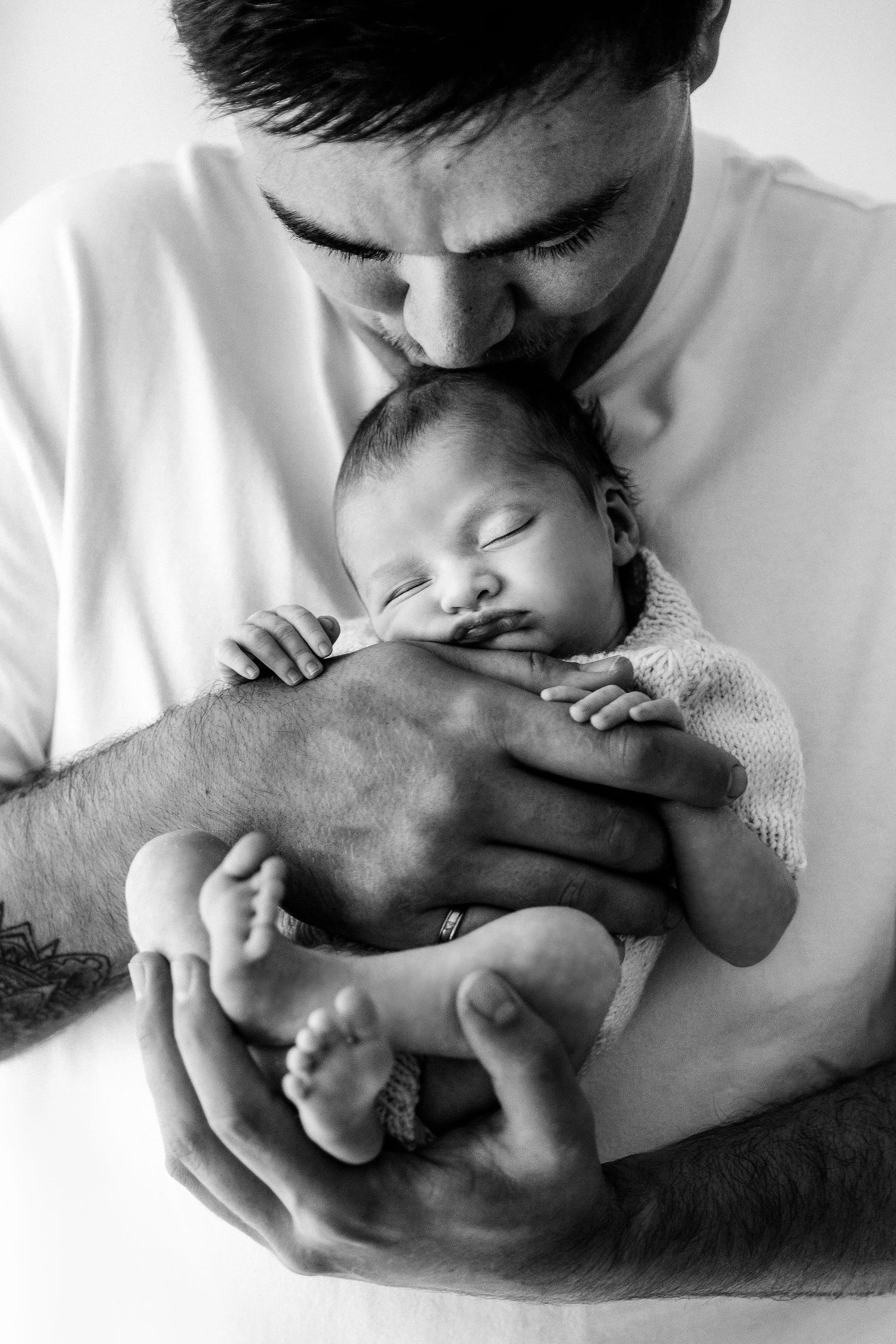 Black and white photo of a father kissing his sleeping swaddled newborn baby.