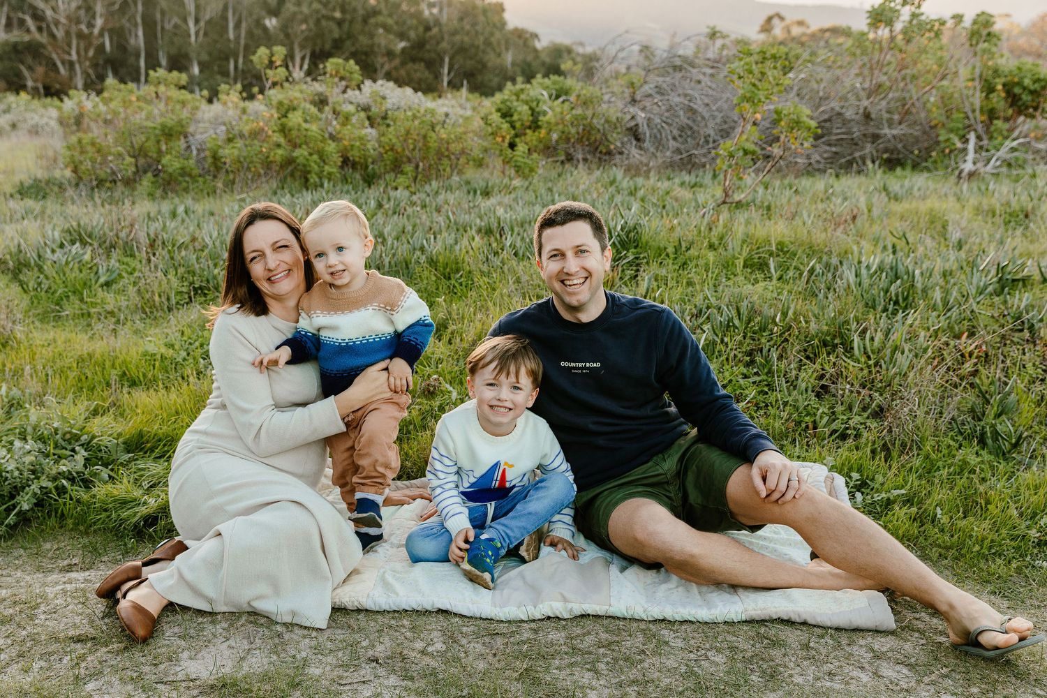 Happy family with two young boys sitting together on a blanket in a green meadow.