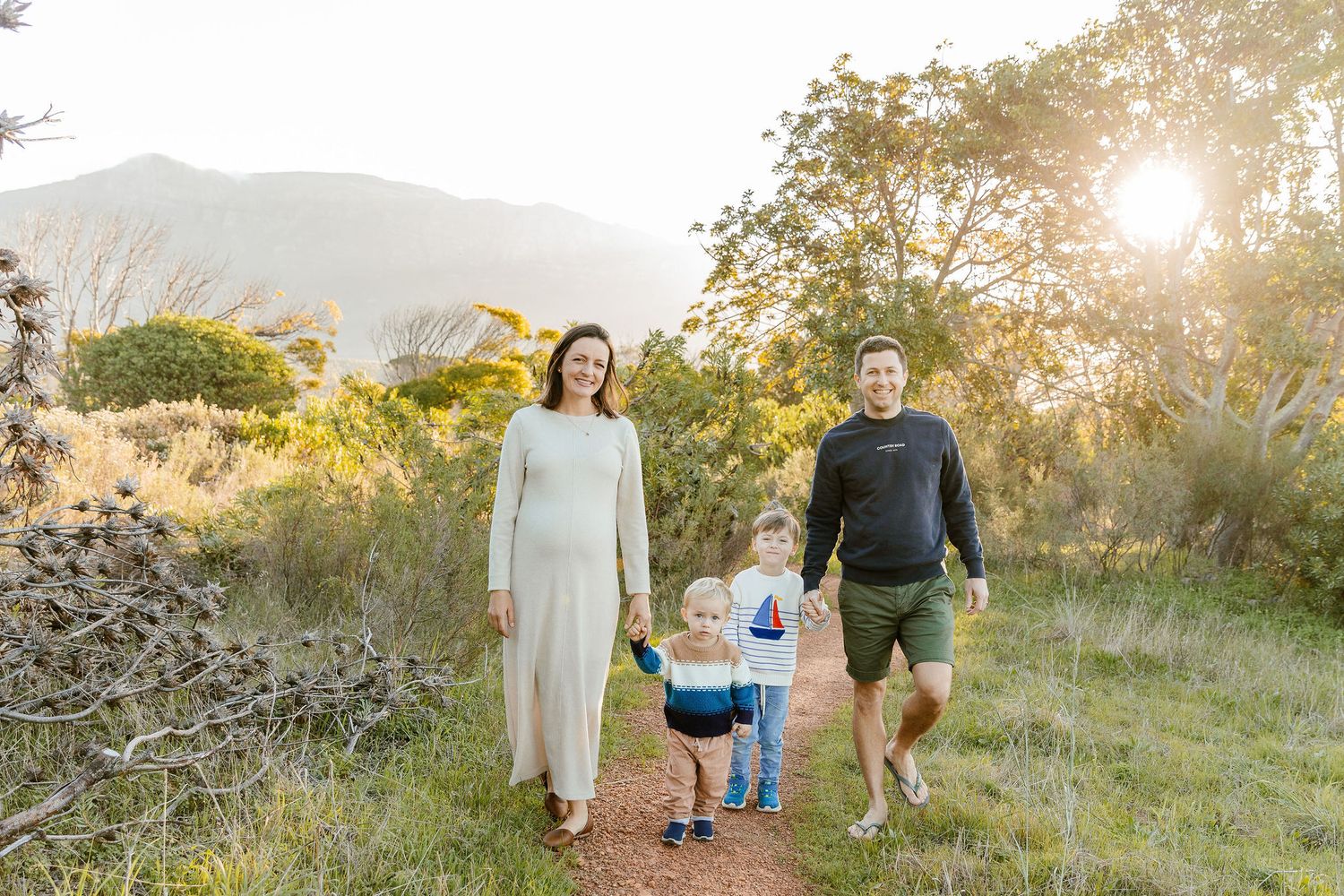 Happy family of four walking together on sunny nature path with mountain backdrop.
