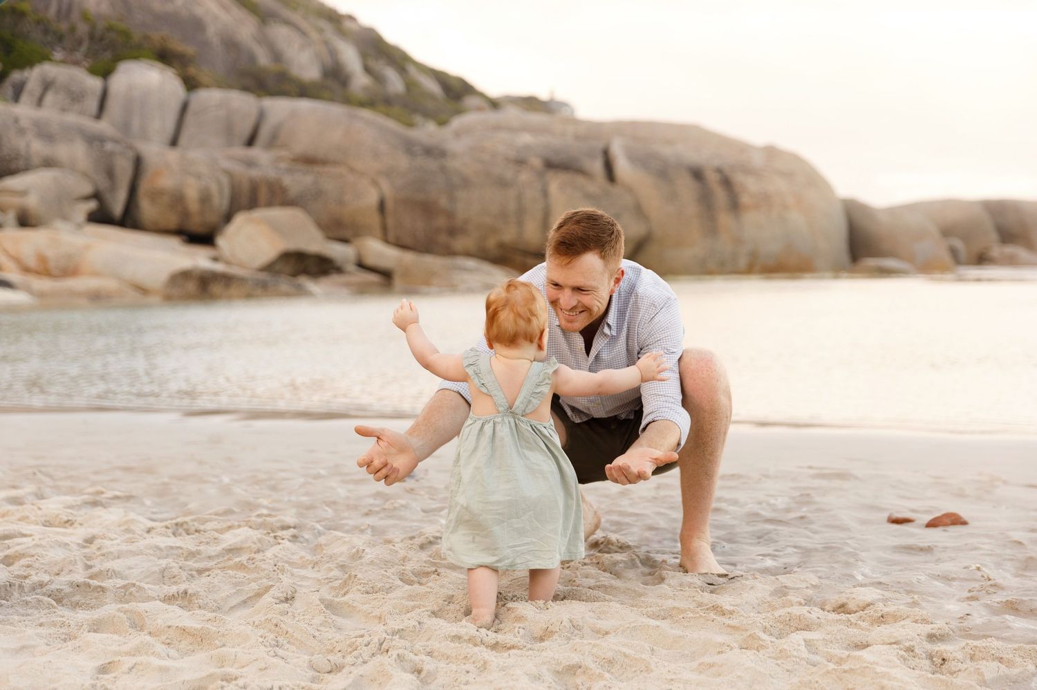 Father crouching on beach sand playing with toddler in green dress.