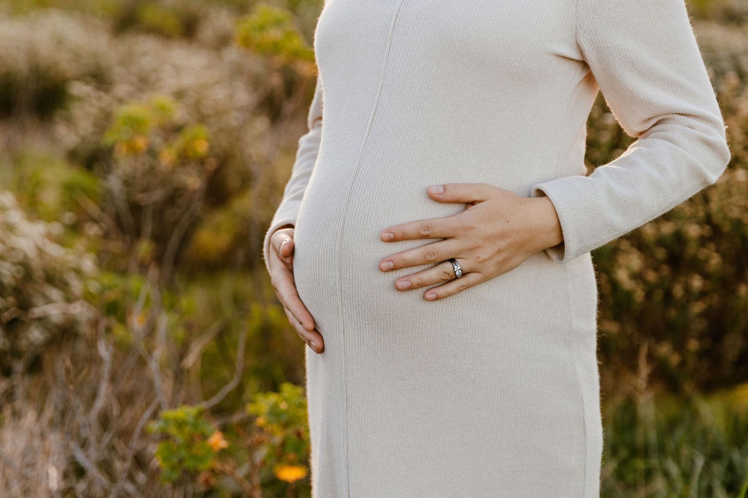 Pregnant woman in cream dress cradling baby bump outdoors with natural foliage background.