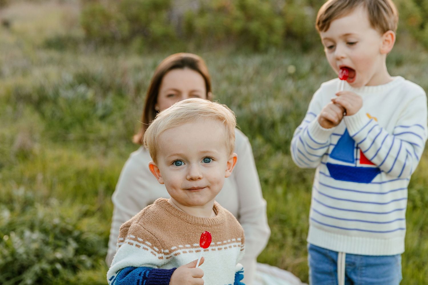 Two young brothers enjoy snacks outdoors while their mother sits behind them in a natural grassy setting.