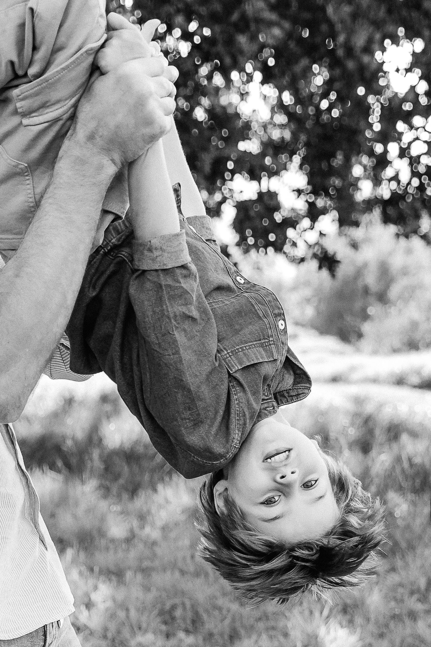 Black and white photo of a child hanging upside down outdoors, held by an adult's hands, smiling joyfully.