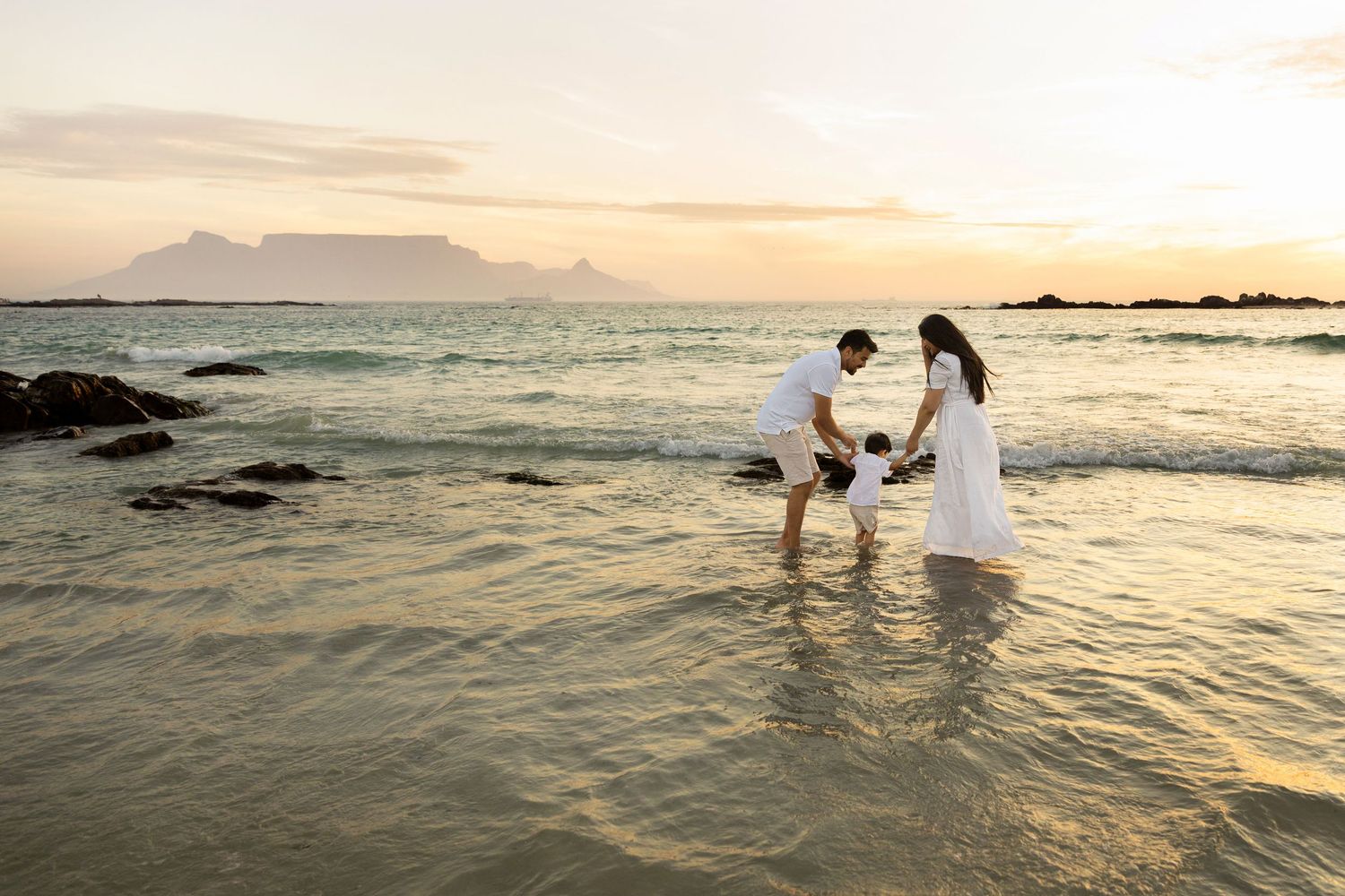 Couple in white outfits playing with child in shallow ocean water at sunset with mountain backdrop.