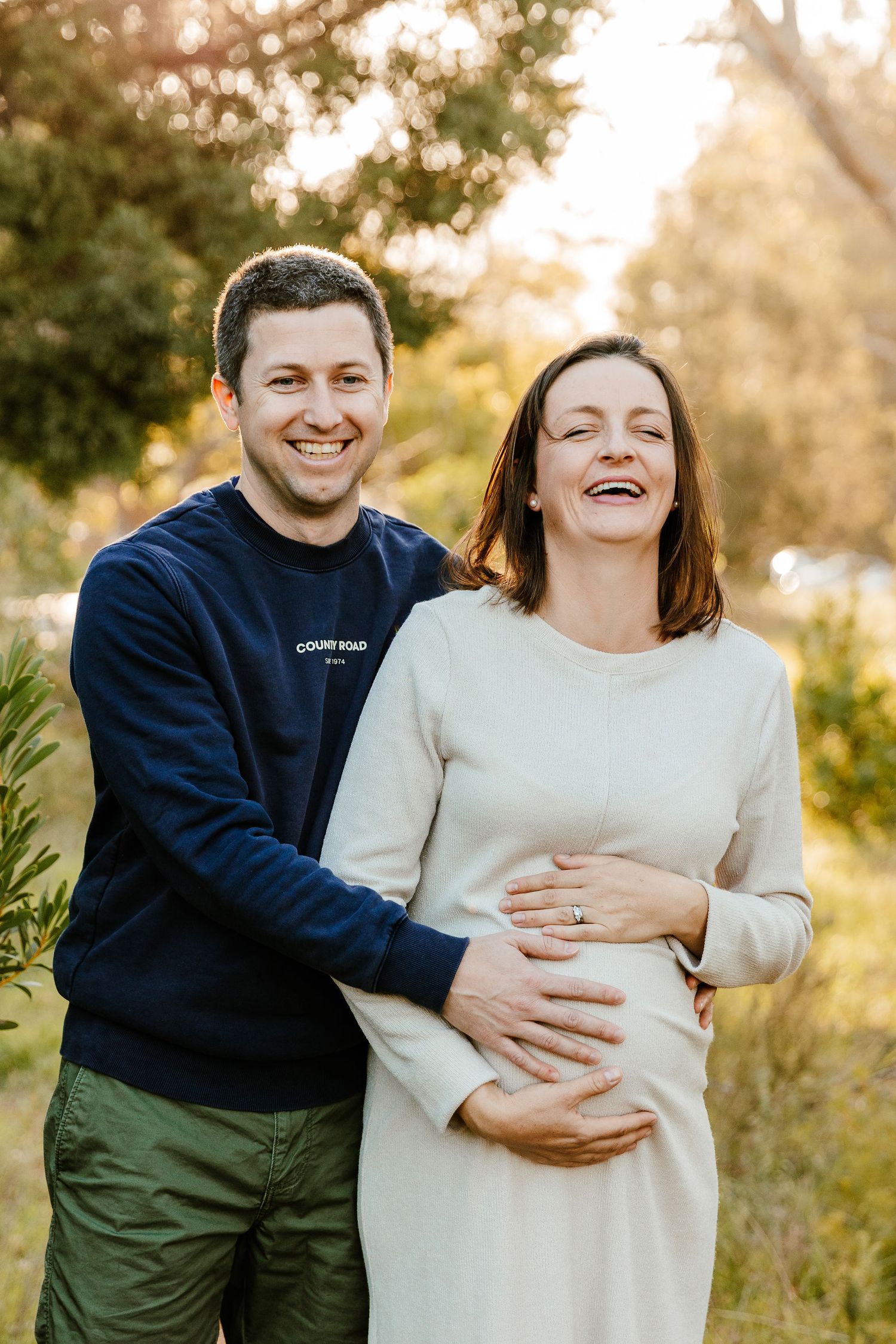 Smiling couple embracing outdoors, pregnant woman in cream dress with partner in navy sweatshirt, golden hour park setting.