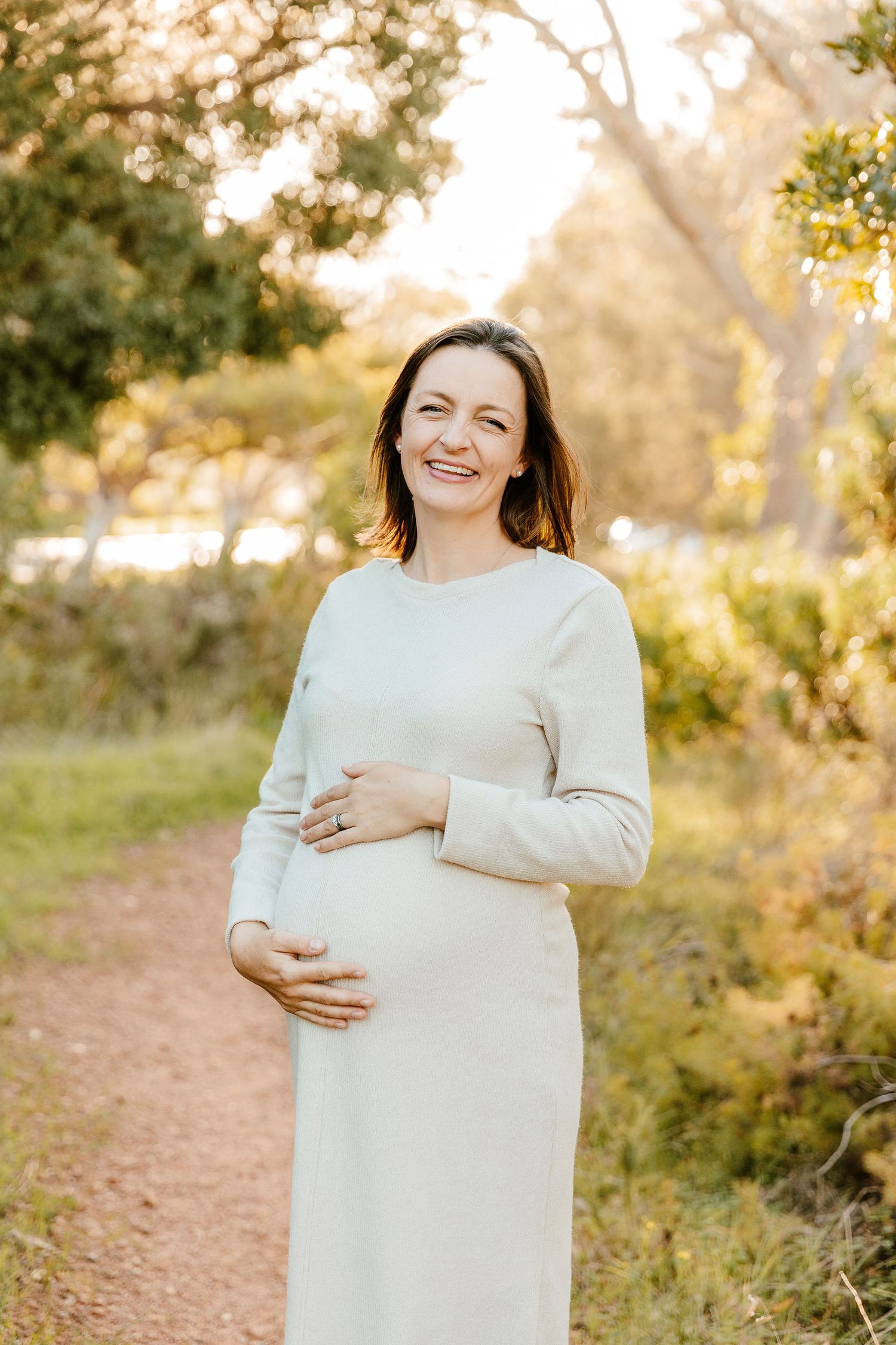 Smiling pregnant woman in white dress cradling baby bump outdoors on a sunlit garden path.