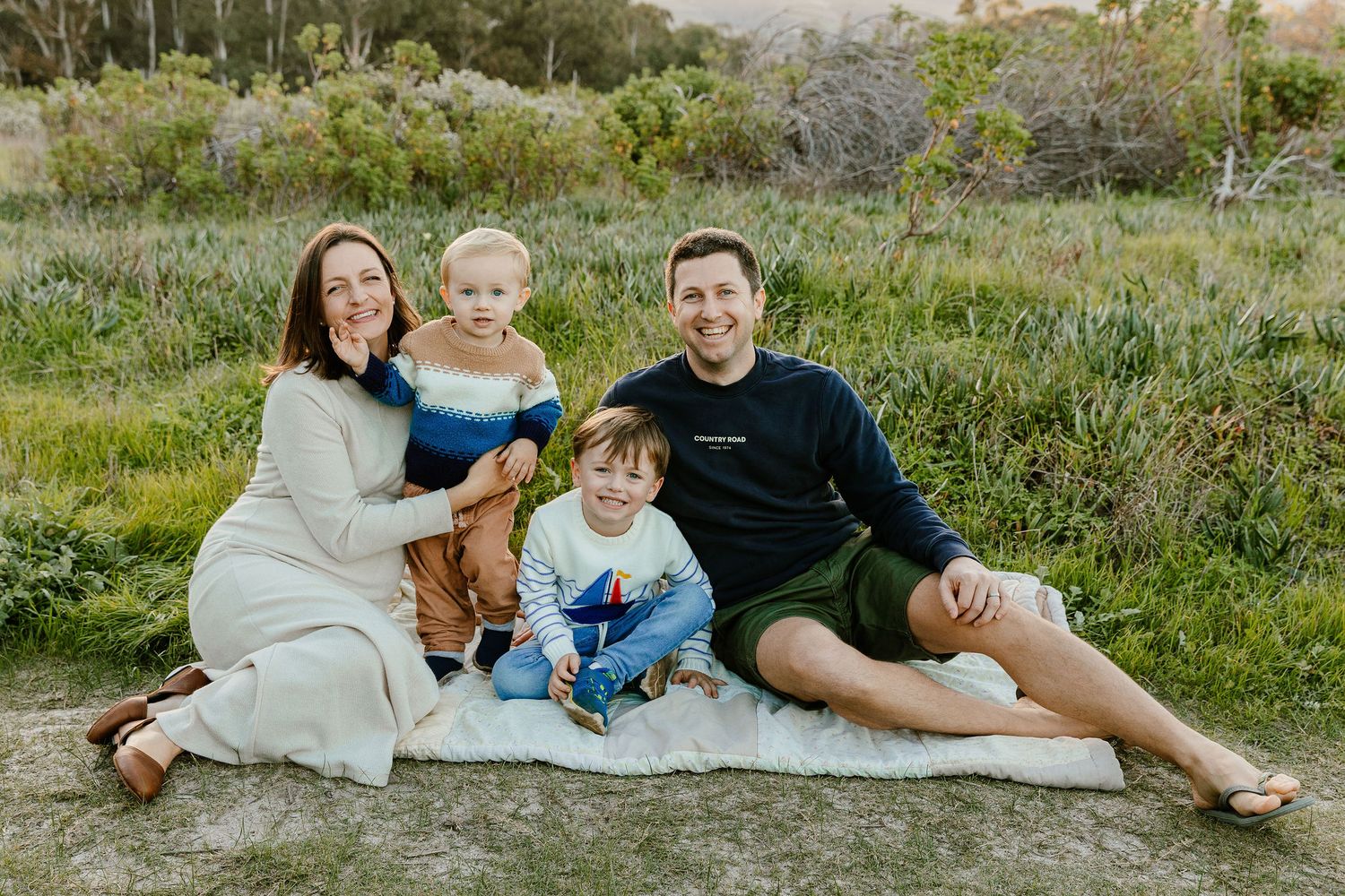Happy family of four sitting on a blanket in a green meadow during golden hour.